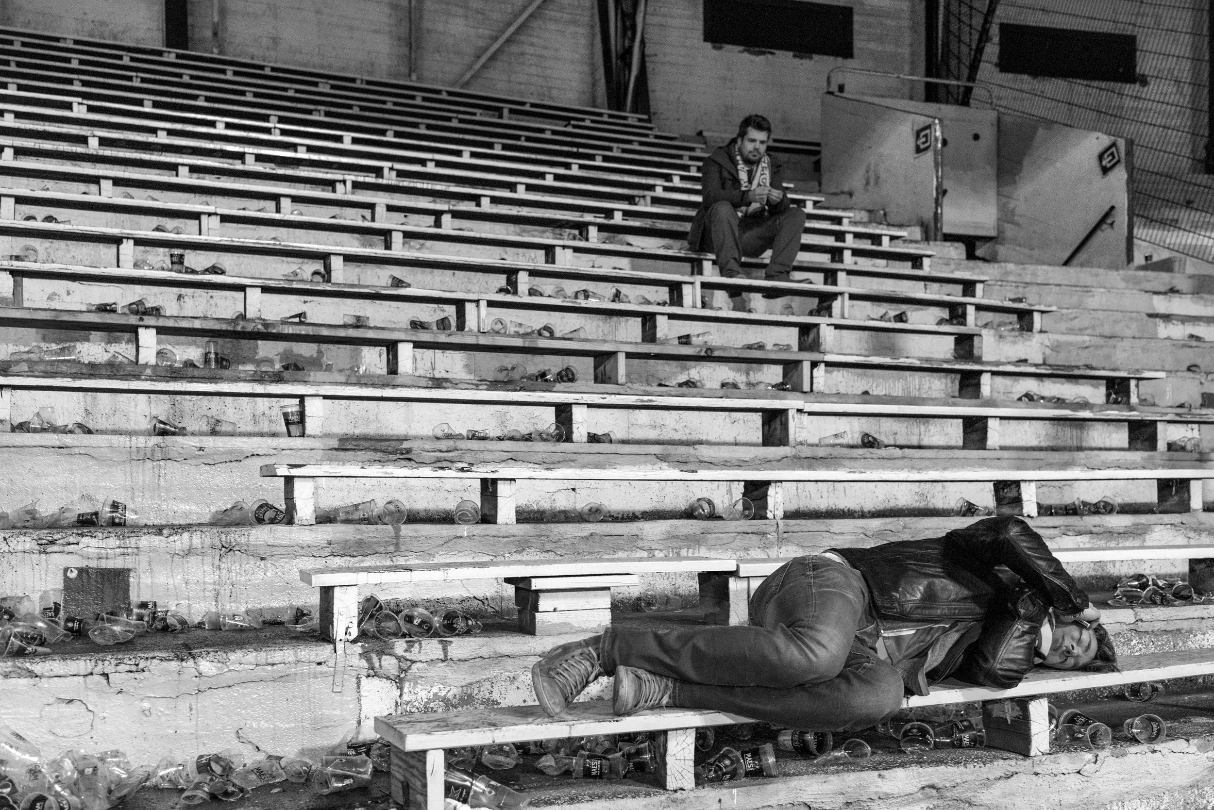 Een man ligt op een bankje in een stadion, terwijl een andere man bovenaan op de tribune zit en op zijn telefoon kijkt, allebei omringd door rommel van lege drankflesjes op de stappen.