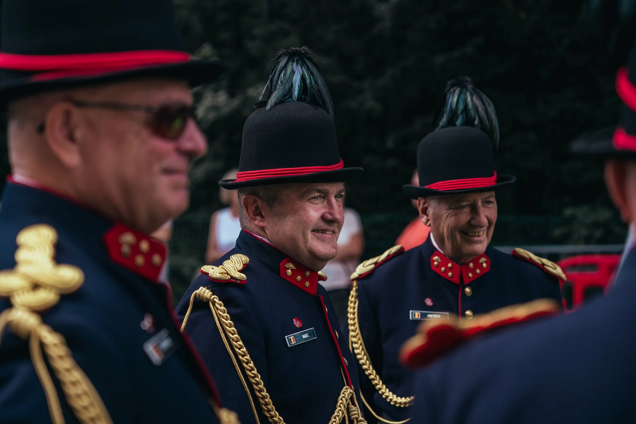 Militairen in uniform en hoge hoeden met veren, lachend en in gesprek, buiten op een bijeenkomst.