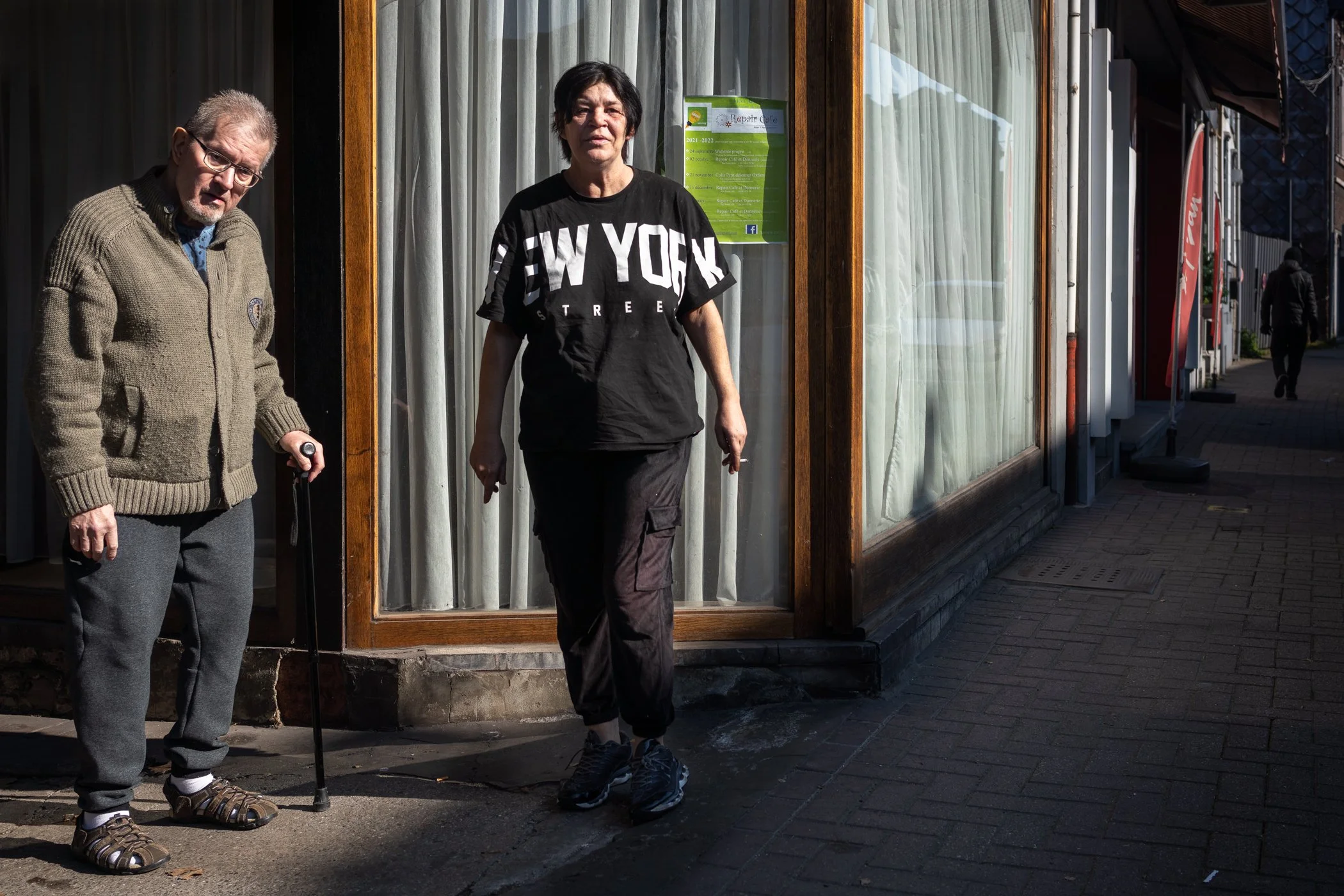 Twee mensen staan buiten een winkel met grote ramen. Een man met een wandelstok en een vrouw in een zwart T-shirt met 'New York' tekst.