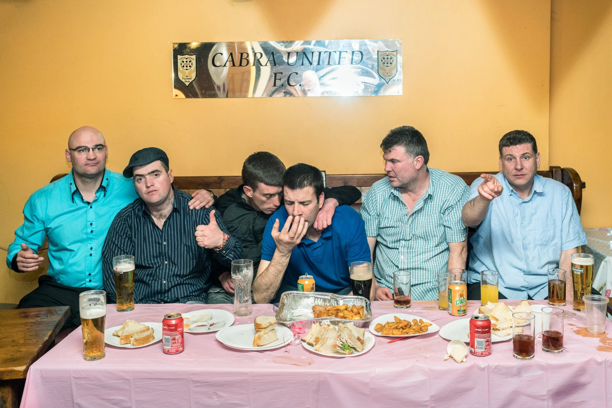 Vijf mannen zitten aan een tafel met eten en drinken, met een spandoek waarop 'CABRA UNITED F.C.' hangt op de muur achter hen.