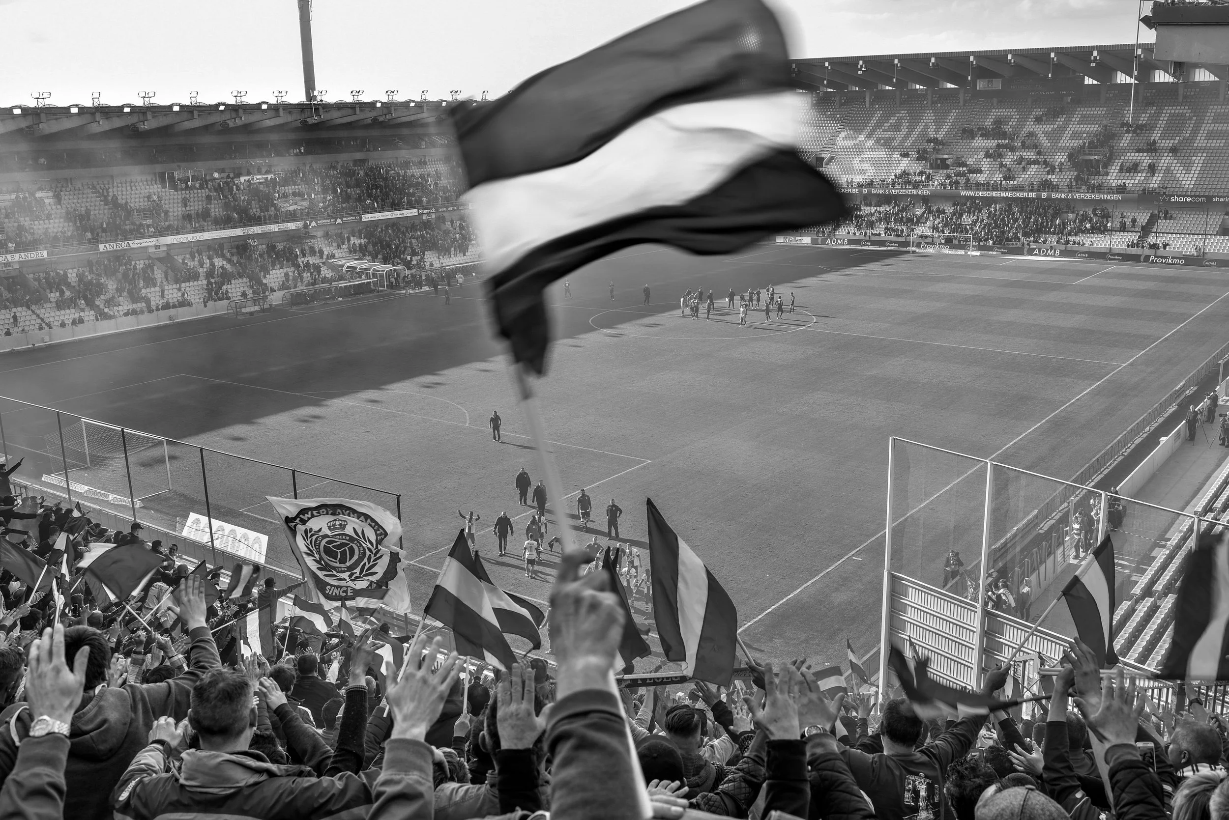 Gezicht op een voetbalstadion met fans die vlaggen zwaaien en spelers op het veld na een wedstrijd, in zwart-wit