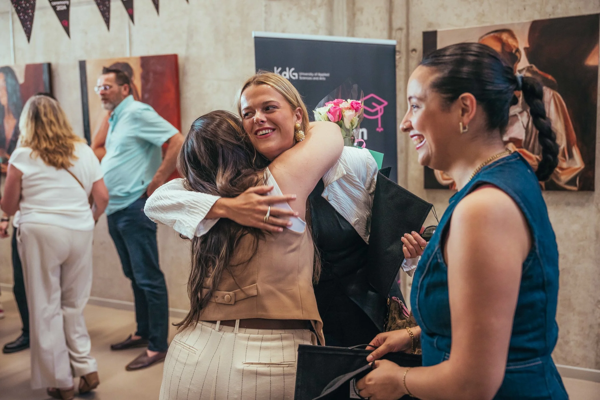 Vrouwen omhelzen elkaar blij in een galerij met kunstwerken aan de muur, glimlachend en genietend van een evenement.