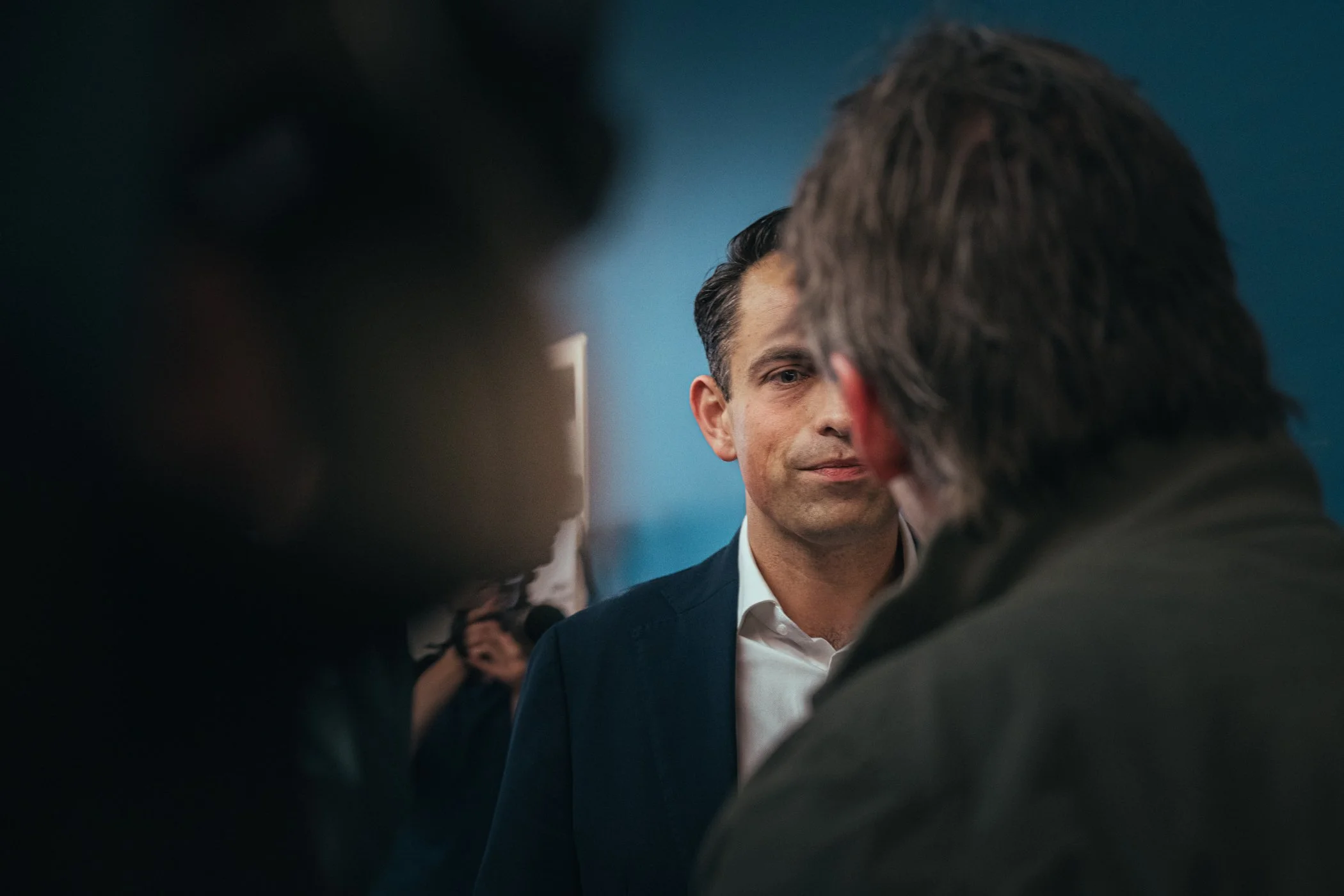 Man in formele kleding kijkt serieus terwijl hij met iemand praat, op de achtergrond andere mensen.