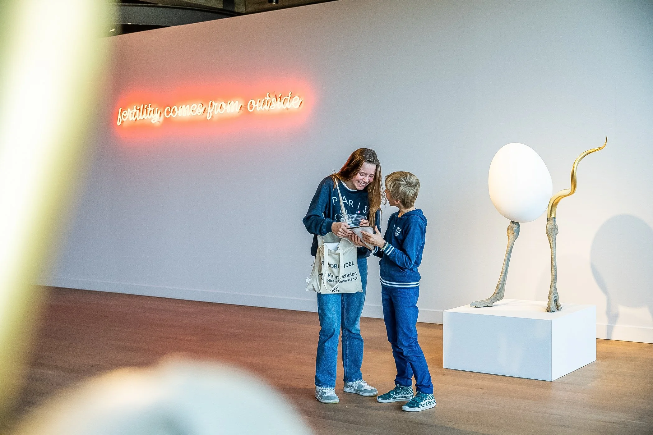 Twee mensen, een vrouw en een jongen, lachen en kijken elkaar aan in een kunstgalerie. Achter hen een witte muur met een neonbord met de tekst 'fertility comes from outside' en een moderne sculptuur van een olifant met een ei op zijn rug.