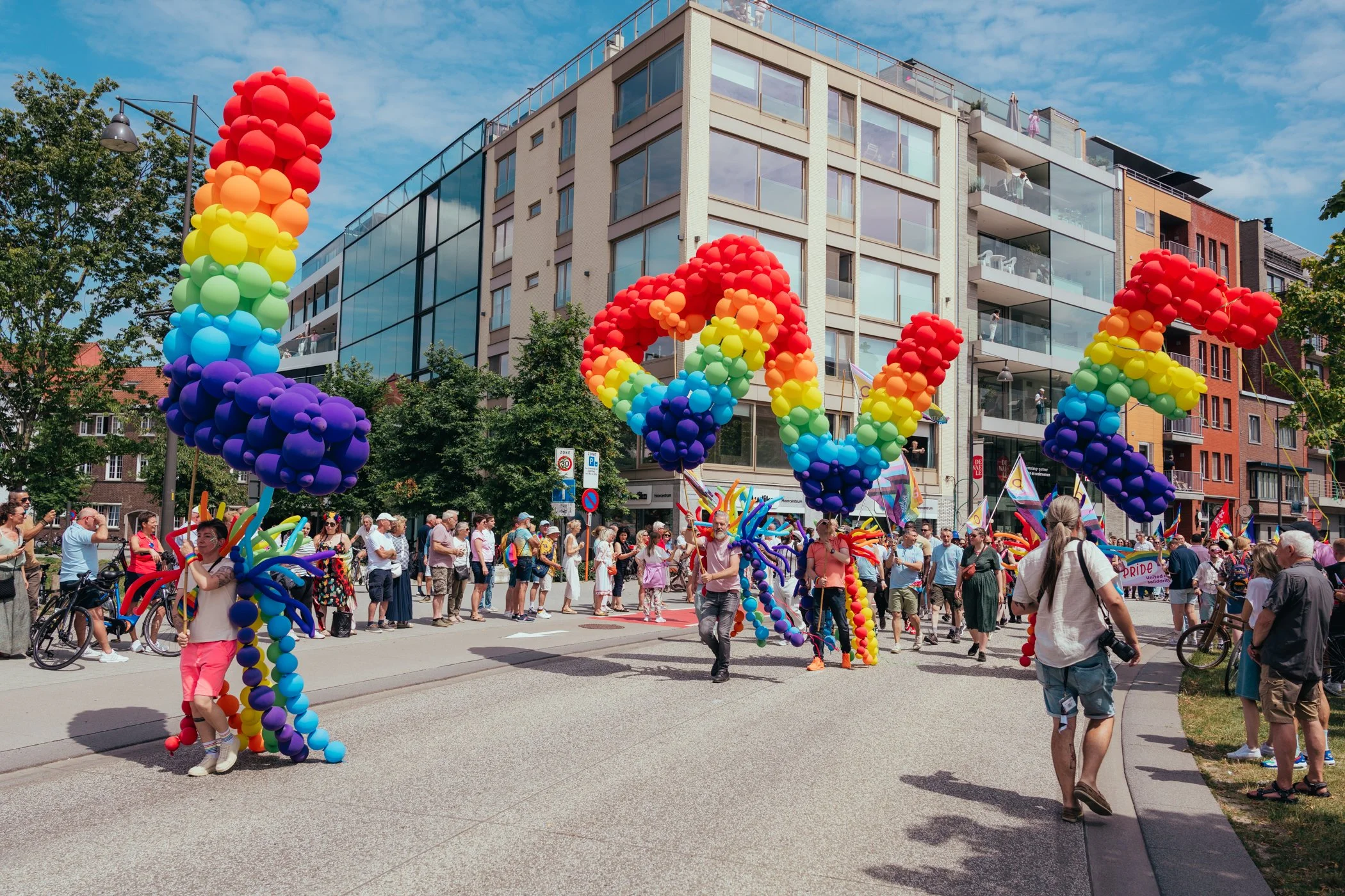 Luchtfoto van een Pride-parade met grote kleurrijke ballonnen die de letters 'LOVE' vormen, omringd door mensen op een straat in een stedelijke omgeving onder een blauwe hemel.