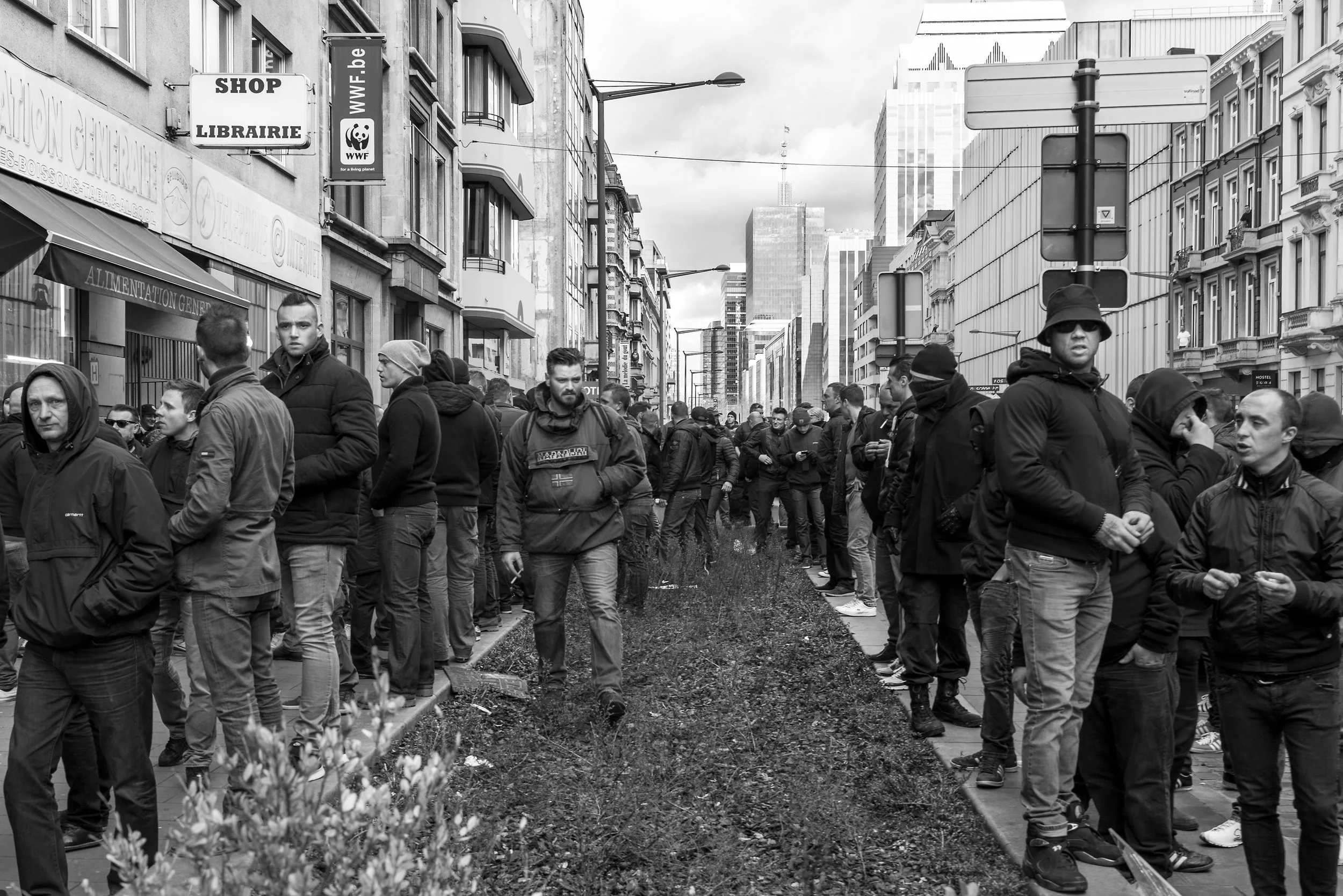 Een menigte mensen staat in een rij langs een straat in een stedelijk gebied, in een zwart-wit foto.