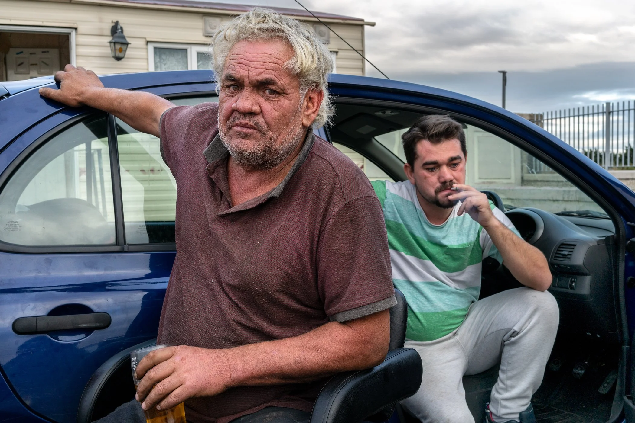 Twee mannen, een oudere man met grijs haar en een paars shirt die in de camera kijkt, en een jongere man met een cigarette en een groene en witte gestreepte shirt die in de auto zit. Ze zijn naast een blauwe auto met een huis op de achtergrond.
