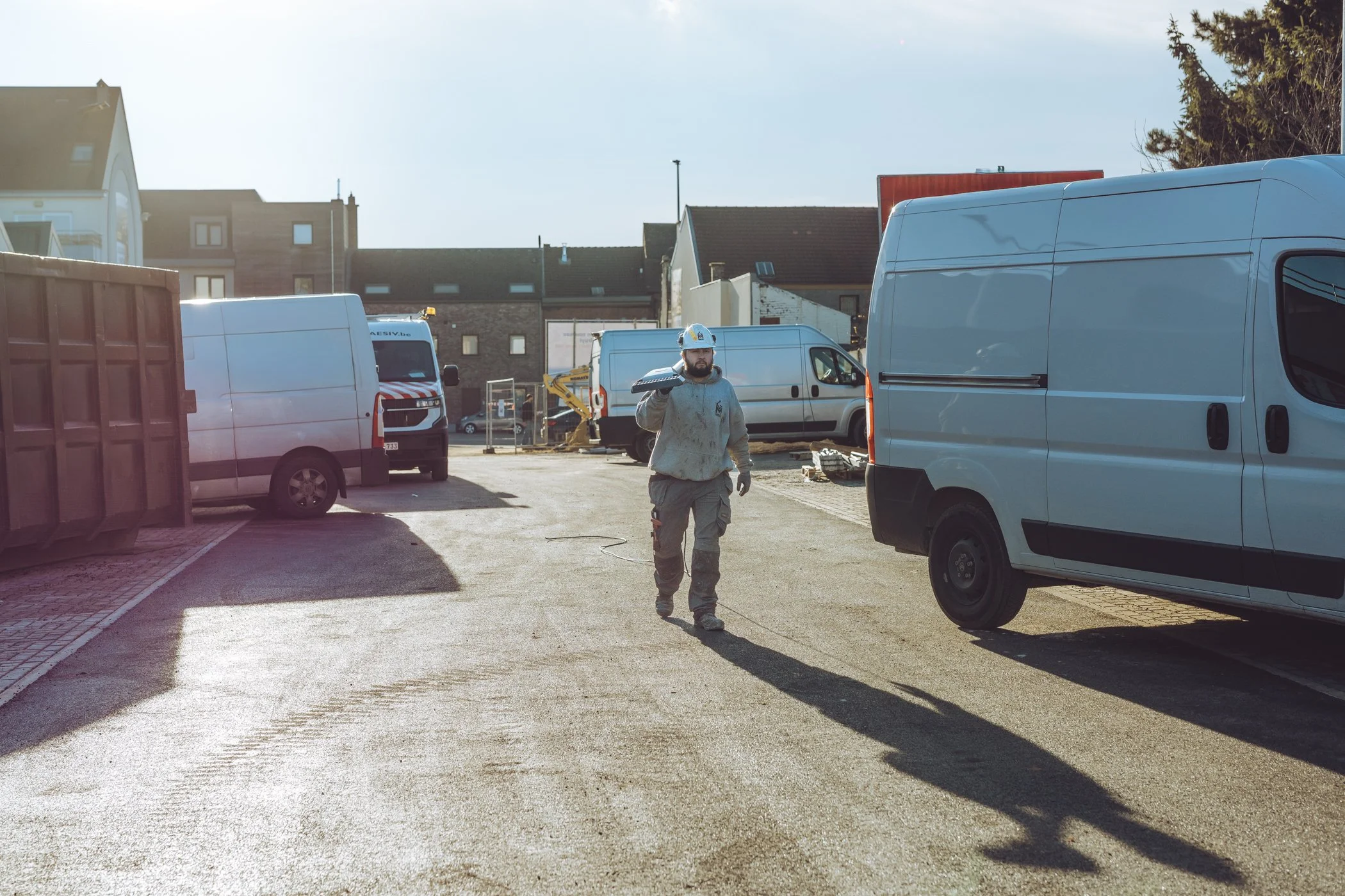 Een bouwvakker in werkkleding en een veiligheidsharnas, lopend in een bouwplaats met meerdere witte bedrijfsvoertuigen en enkele gebouwen op de achtergrond onder een blauwe hemel.