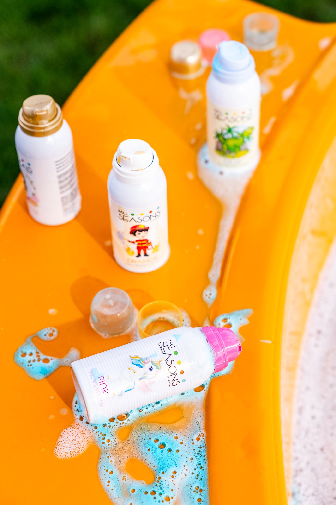 Flesjes zonnebrandcrème en een tube met een unicorn-illustratie liggen op een oranje kinderbadje, water en schuim eromheen, buiten op gras.