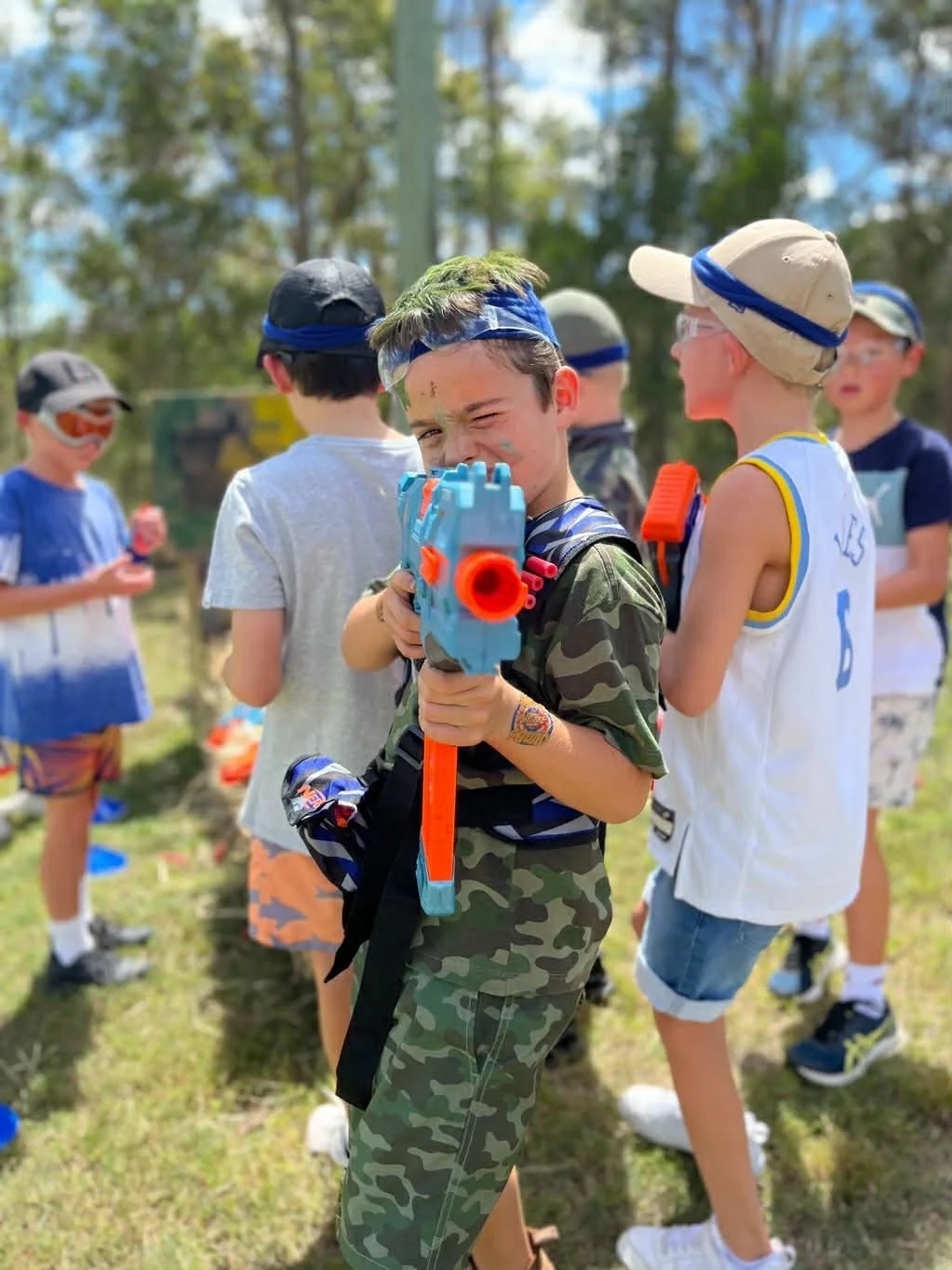 A young boy with a toy laser gun pointing it at the camera, surrounded by other kids outdoors on a sunny day, some wearing hats and sunglasses, with trees in the background.