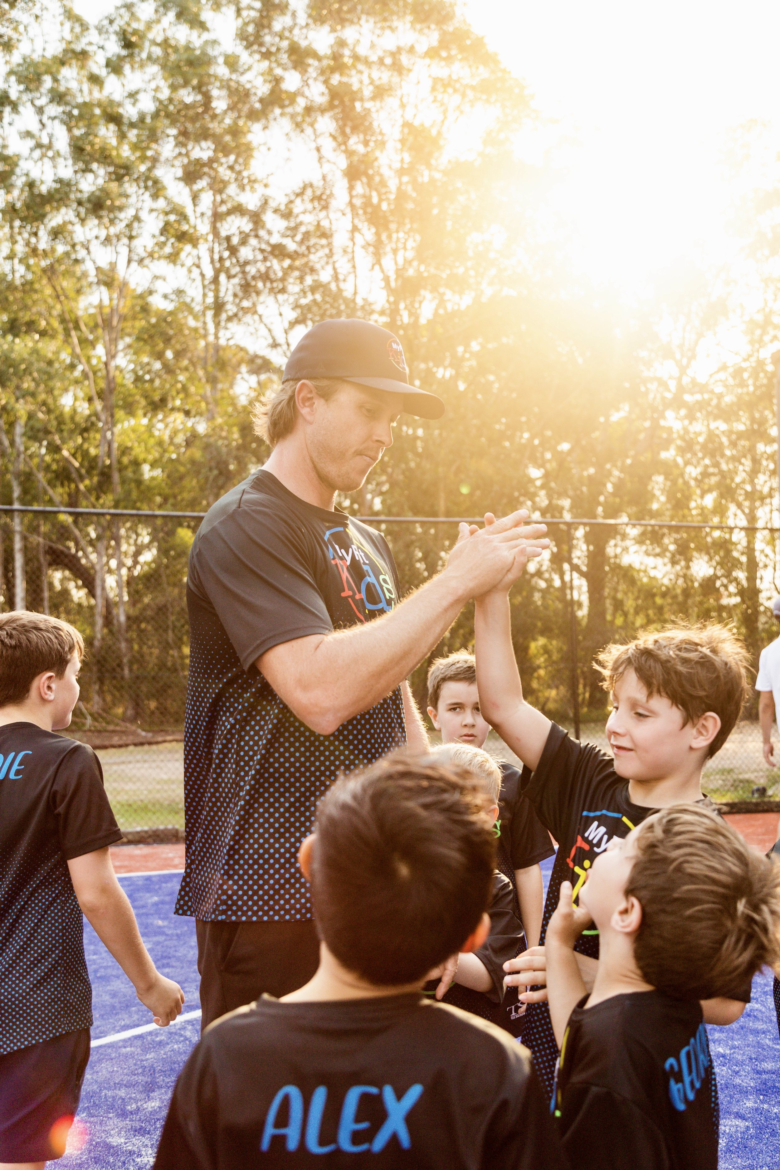 A coach giving a high-five to a young boy during a youth sports practice on an outdoor court at sunset.