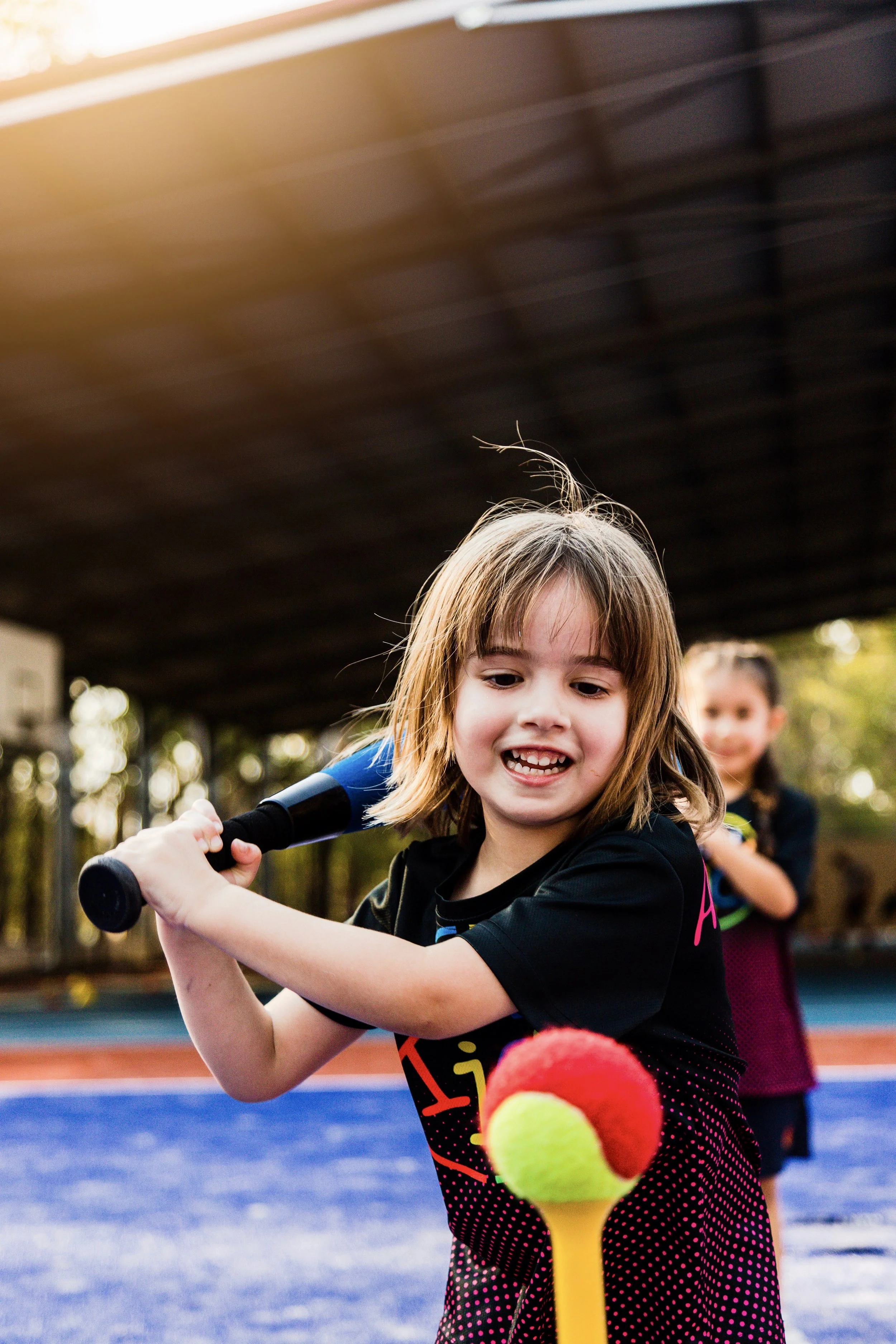 A young girl playing tennis on a blue court, holding a tennis racket behind a large red and yellow tennis ball, with another girl in the background watching.