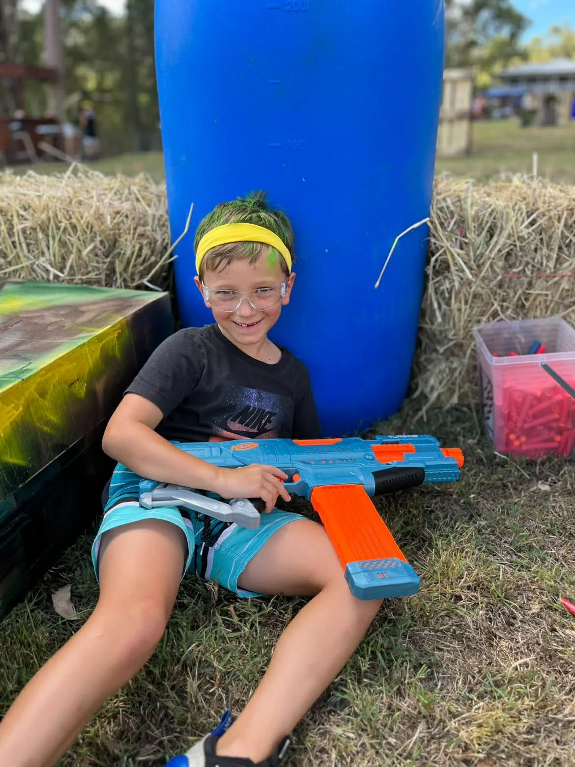 A boy with glasses, wearing a black Nike t-shirt, blue shorts, and a yellow headband, is sitting on the grass next to hay bales. He is smiling and holding a large blue and orange Nerf blaster, with a small toy gun in his lap. There is a large blue barrel behind him and a pink container filled with small toys or parts on his right.