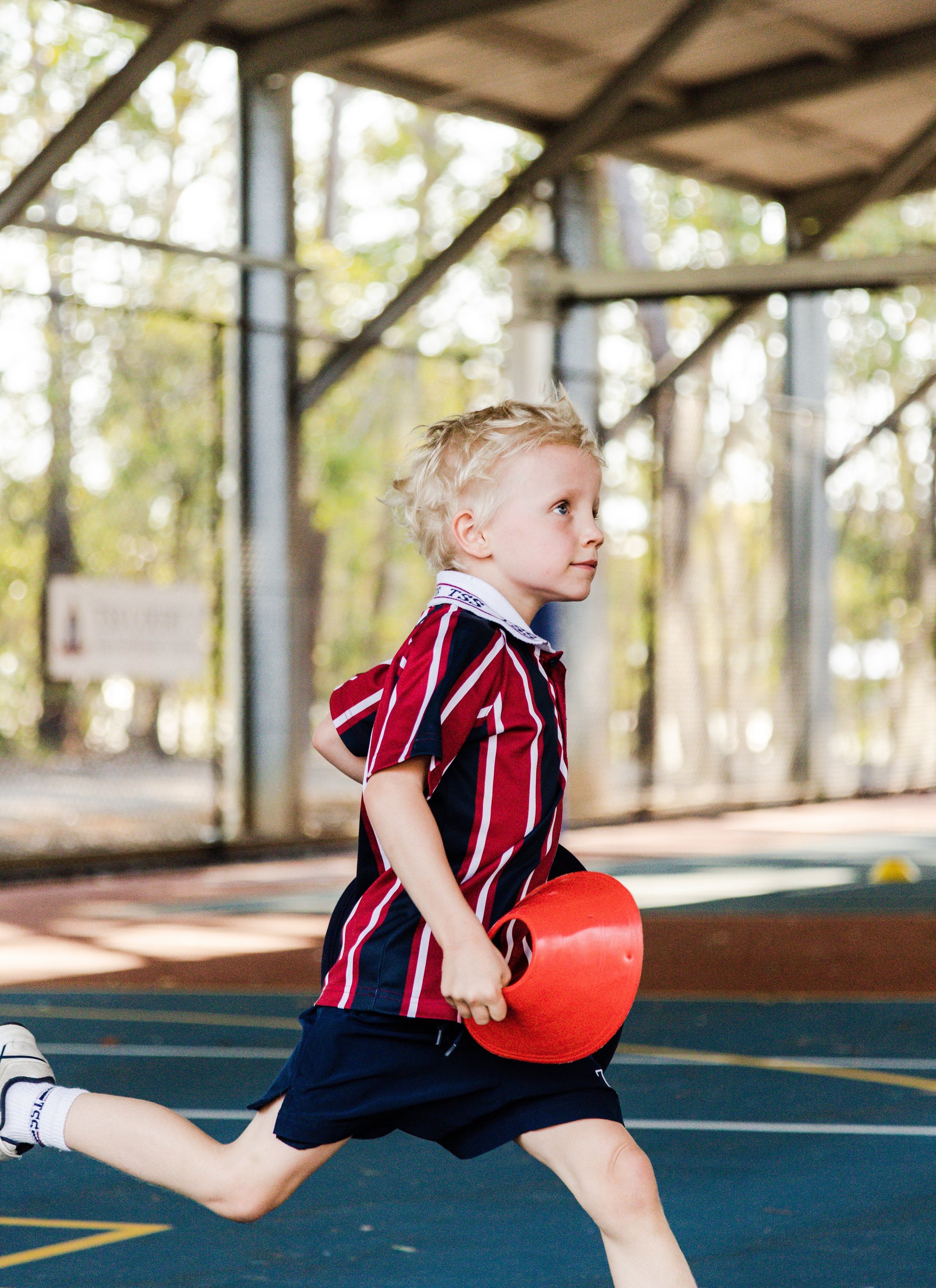 Young boy running on a sports court, holding a red relay baton, under a covered outdoor structure with trees in the background.