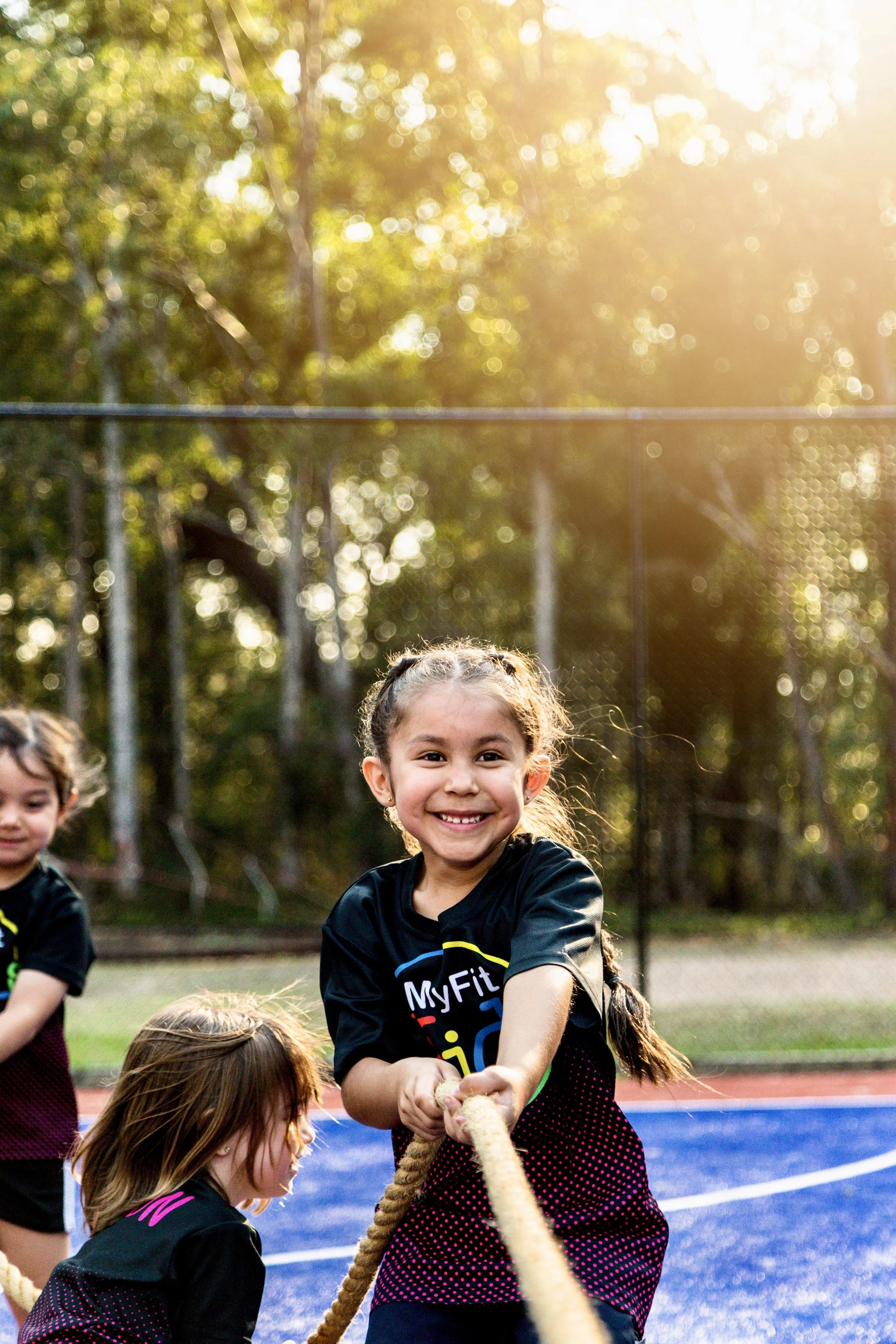Children playing tug of war outdoors on a sunny day, with a girl smiling at the camera in the foreground.