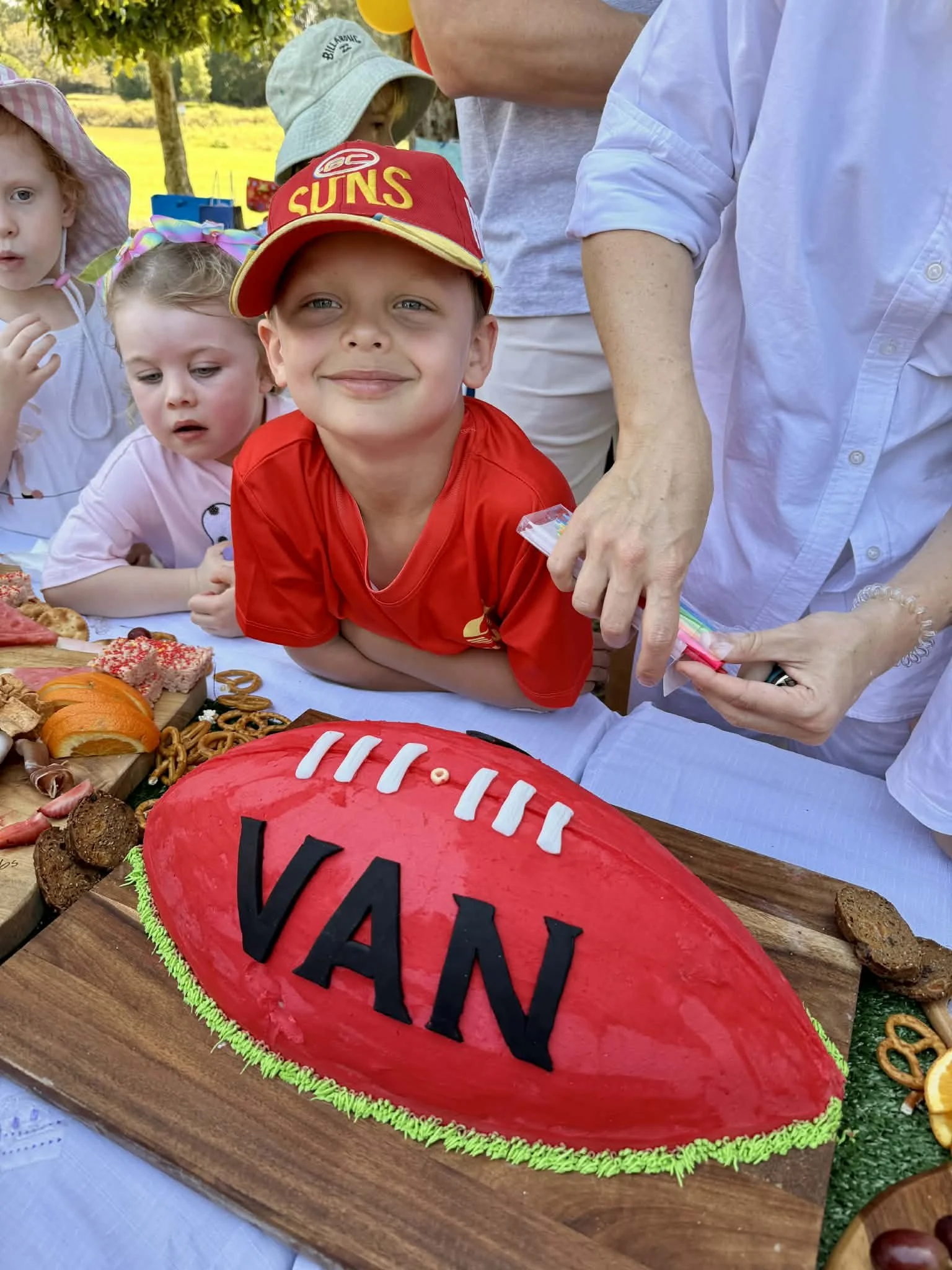A boy in a red shirt and a red baseball cap with the word 'SUNS' is smiling at a sports-themed cake shaped like a football with the name 'VAN' on it. Several children and adults are gathered around the table outdoors with food and decorations.