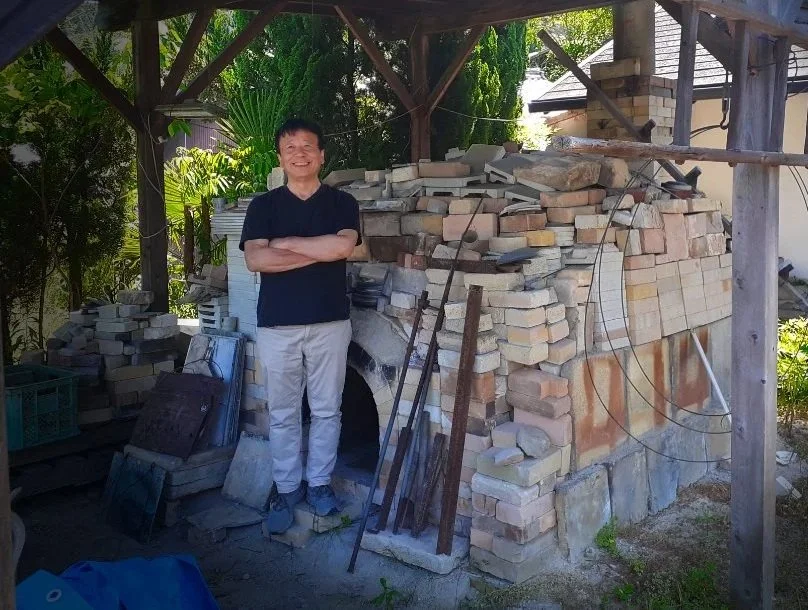 Potter Ikuhiko Shibata standing with crossed arms and smiling next to a brick oven outdoors.
