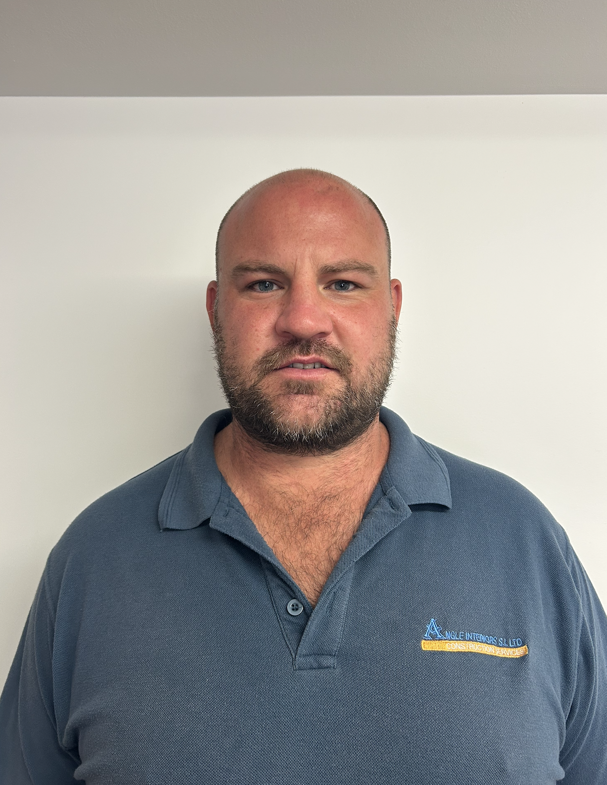 A woman with short blonde hair, wearing a sleeveless orange top and beige pants, sitting on a beige bench, writing in a notebook. Sunlight casts a shadow of a plant on the white wall beside her, and a large green potted plant is to her left.