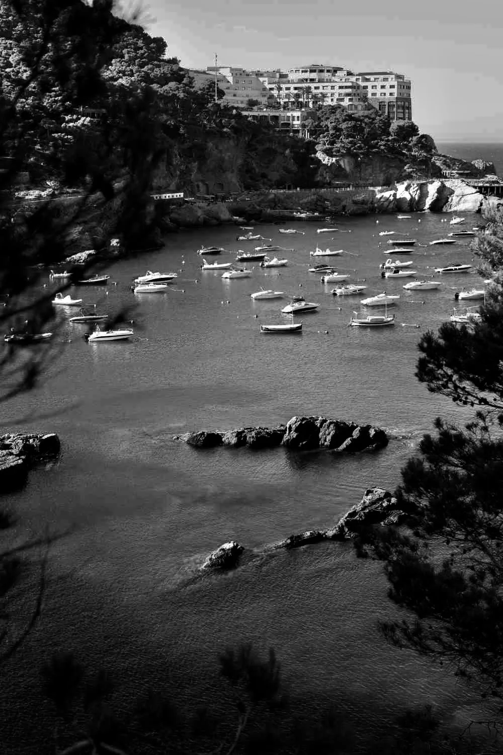 A coastal scene with boats anchored in a calm bay, surrounded by rocky cliffs, trees, and a hillside with large buildings overlooking the water.