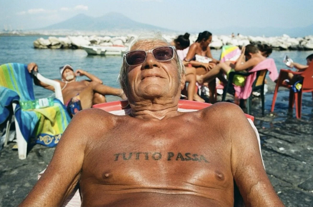 An elderly man with gray hair, sunglasses, and a tattoo on his chest reading "TUTTO PASSA" relaxing outdoors near the water with other people lounging and sitting on chairs, with mountains in the background.