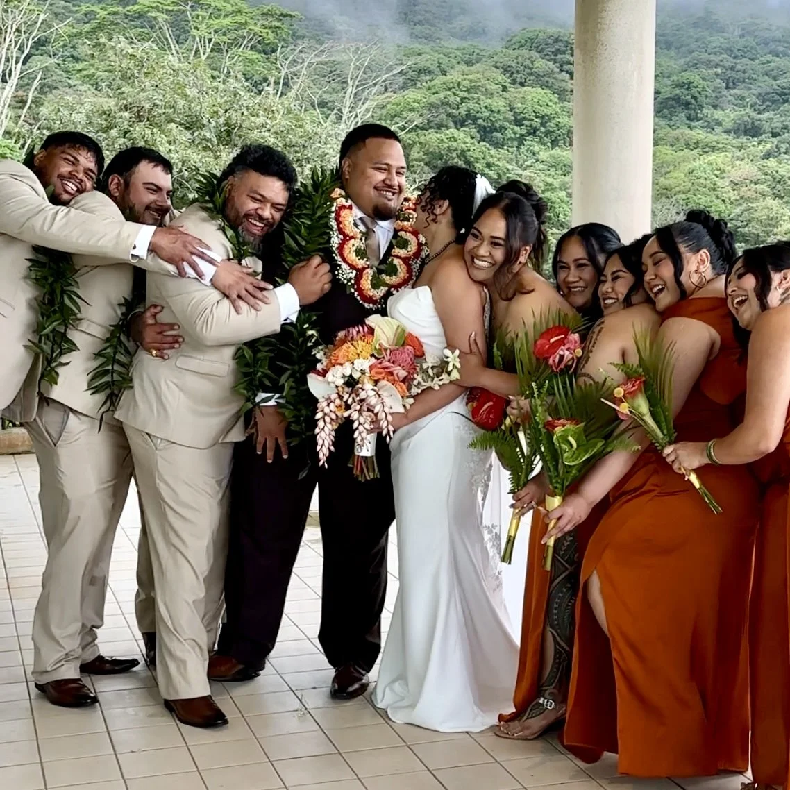 Entire wedding party surrounding and hugging bride and groom in joyful celebration at Ko'olau Ballrooms Oahu