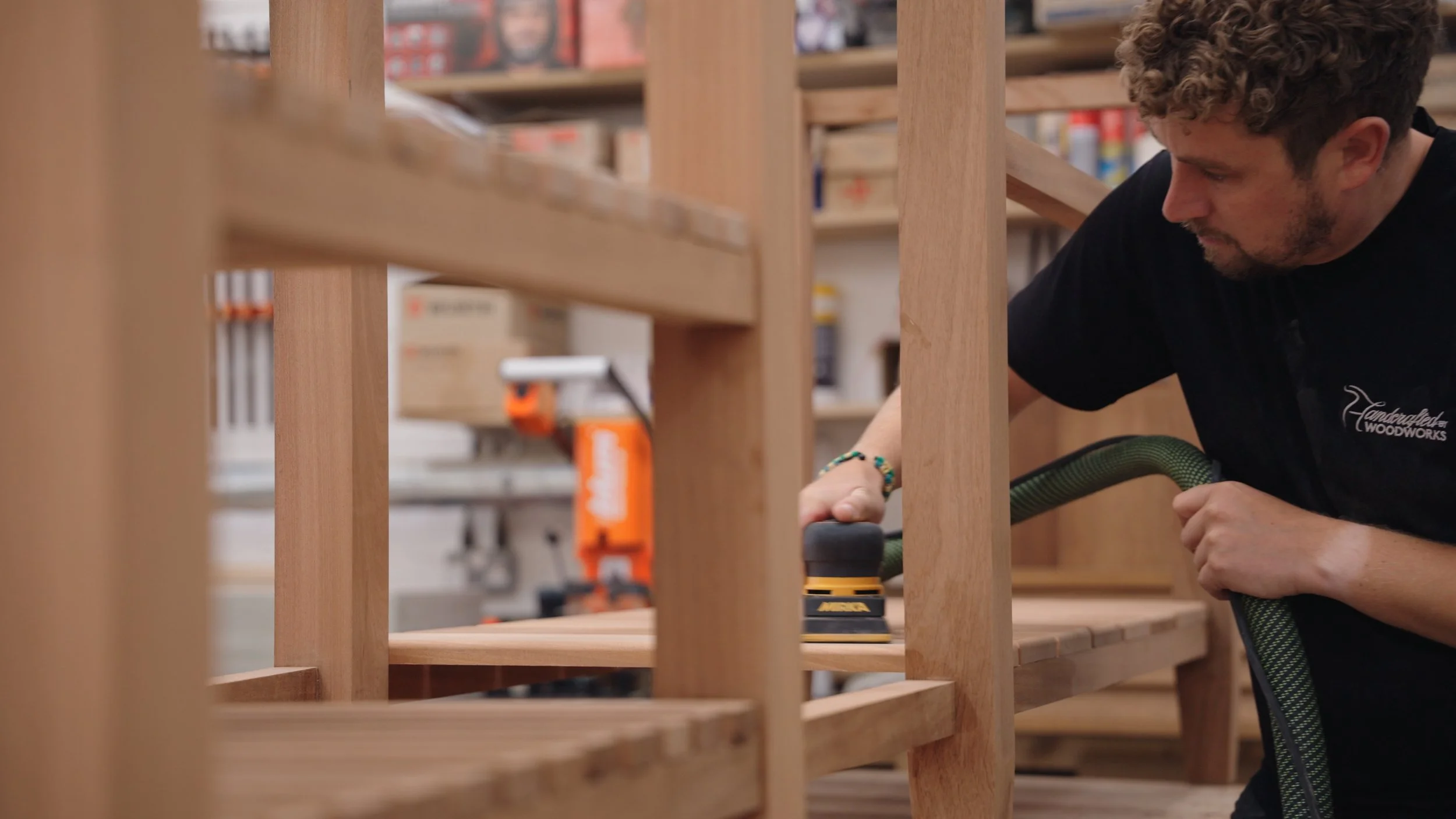 A man working on a wooden furniture piece in a woodworking shop, using a power sander.