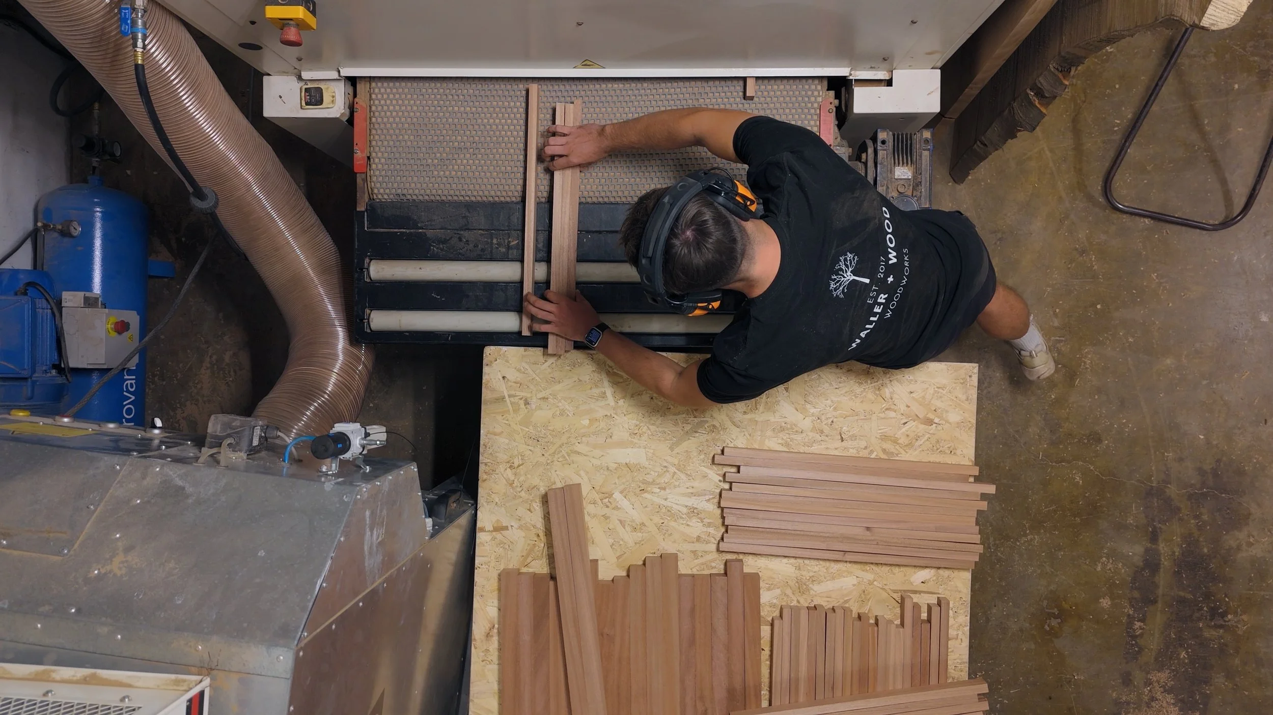 A man wearing a black t-shirt, headphones, and a beanie is working at a woodworking machine, sanding or shaping wood pieces. There are various wooden strips and planks on a work surface in front of him. The workshop contains air ducts, a blue air com