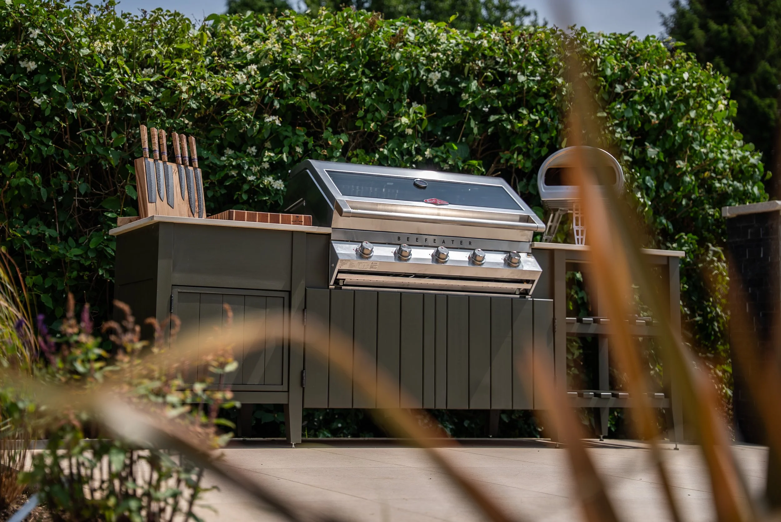 Outdoor kitchen setup with a stainless steel BBQ grill on a wooden cabinet, surrounded by lush green foliage.