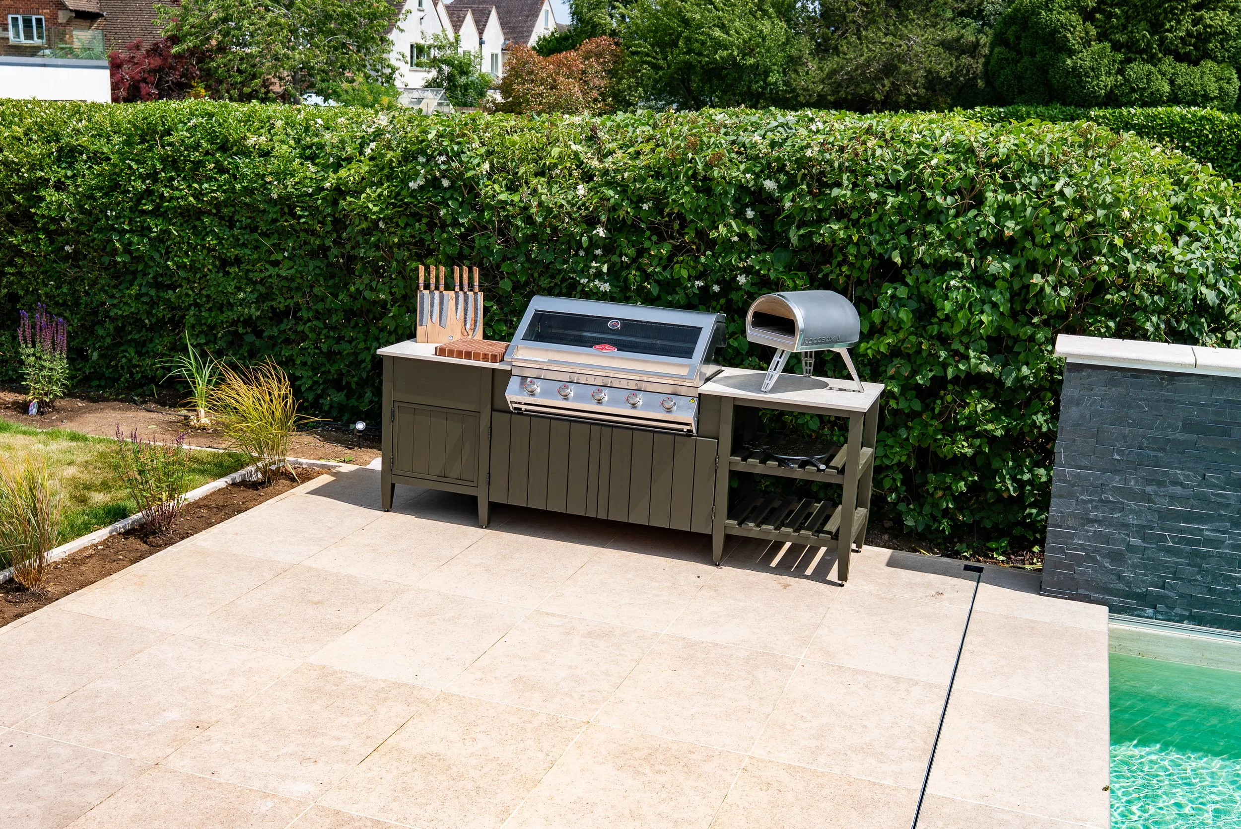 Outdoor patio with a grill and pizza oven next to a garden with small plants, surrounded by a tall green hedge and residential houses in the background.