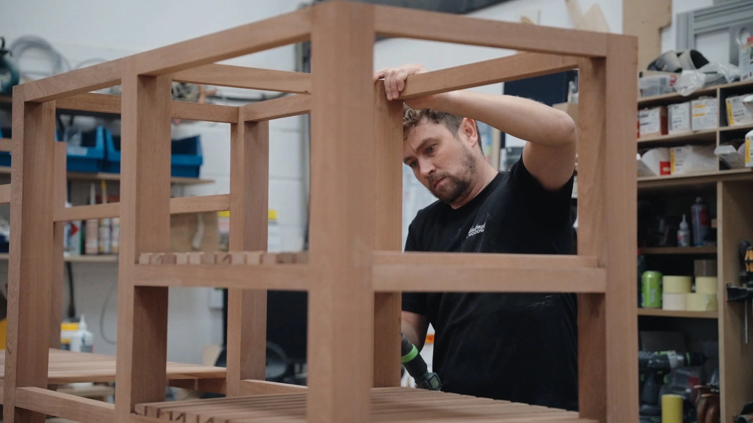 A man assembling a wooden frame structure in a workshop, with shelves of tools and supplies in the background.