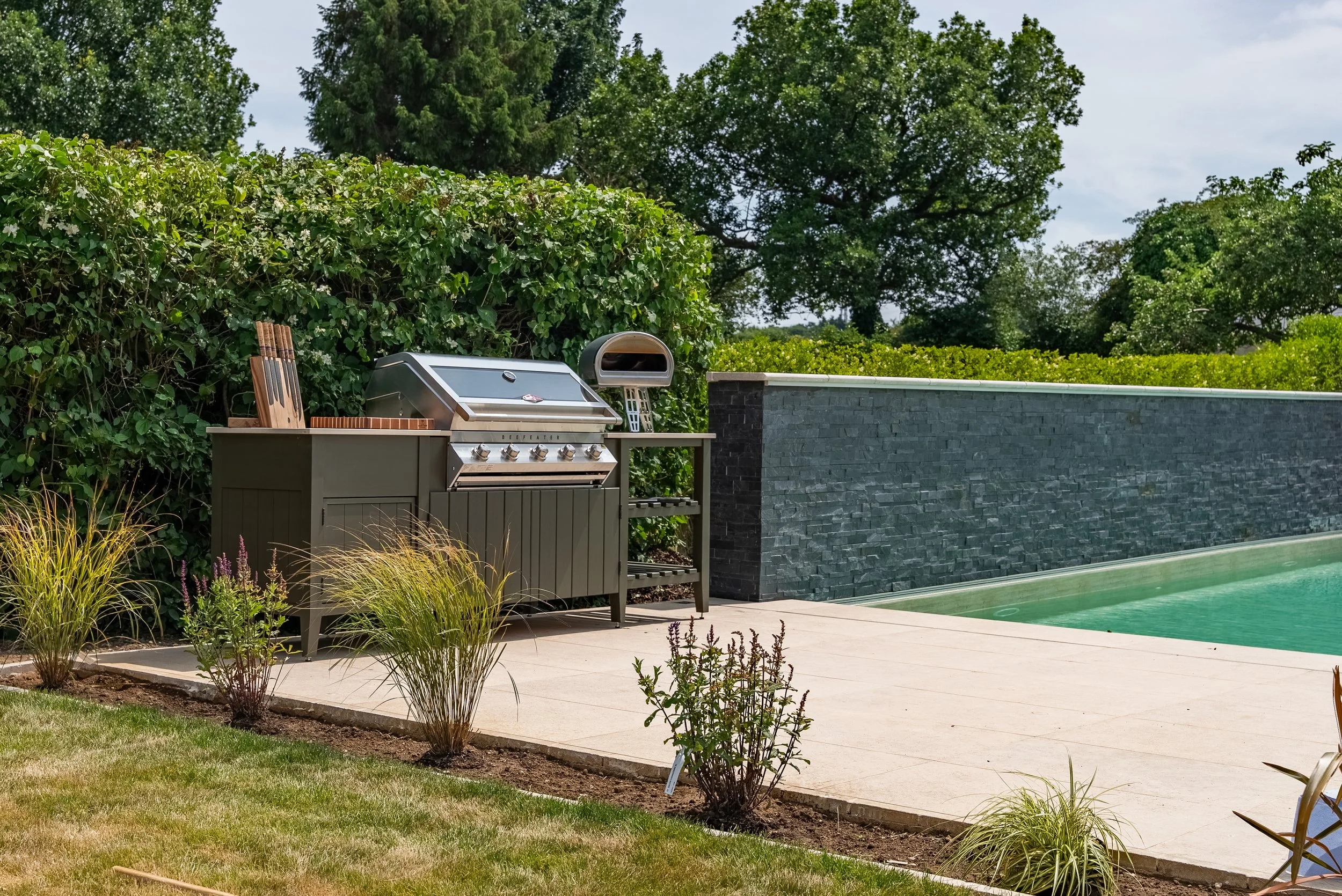 Outdoor swimming pool with a gray barbecue grill and surrounding plants, hedge, and trees.
