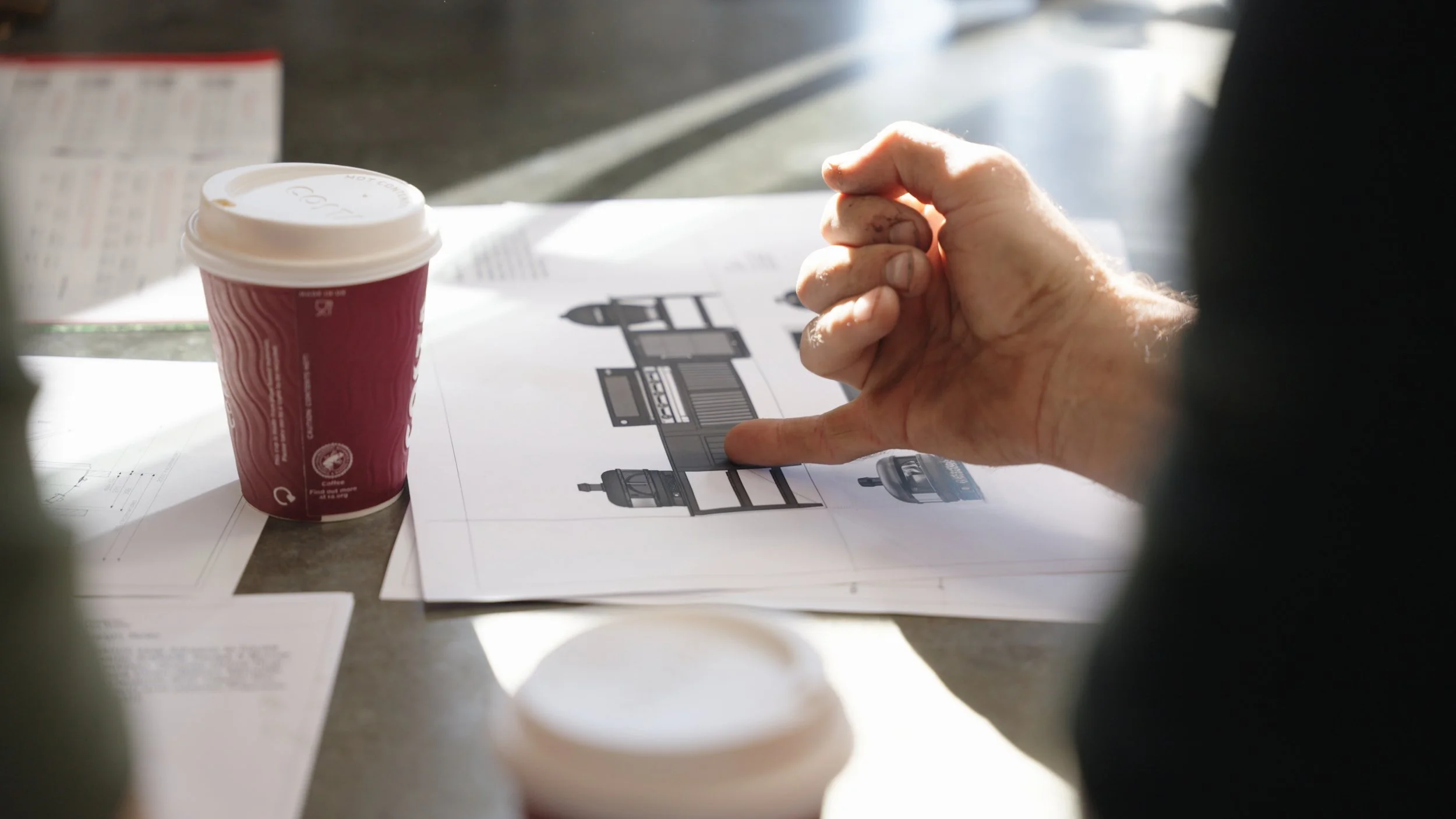 Person pointing at a design plan placed on a table, with a coffee cup nearby, in a well-lit environment.