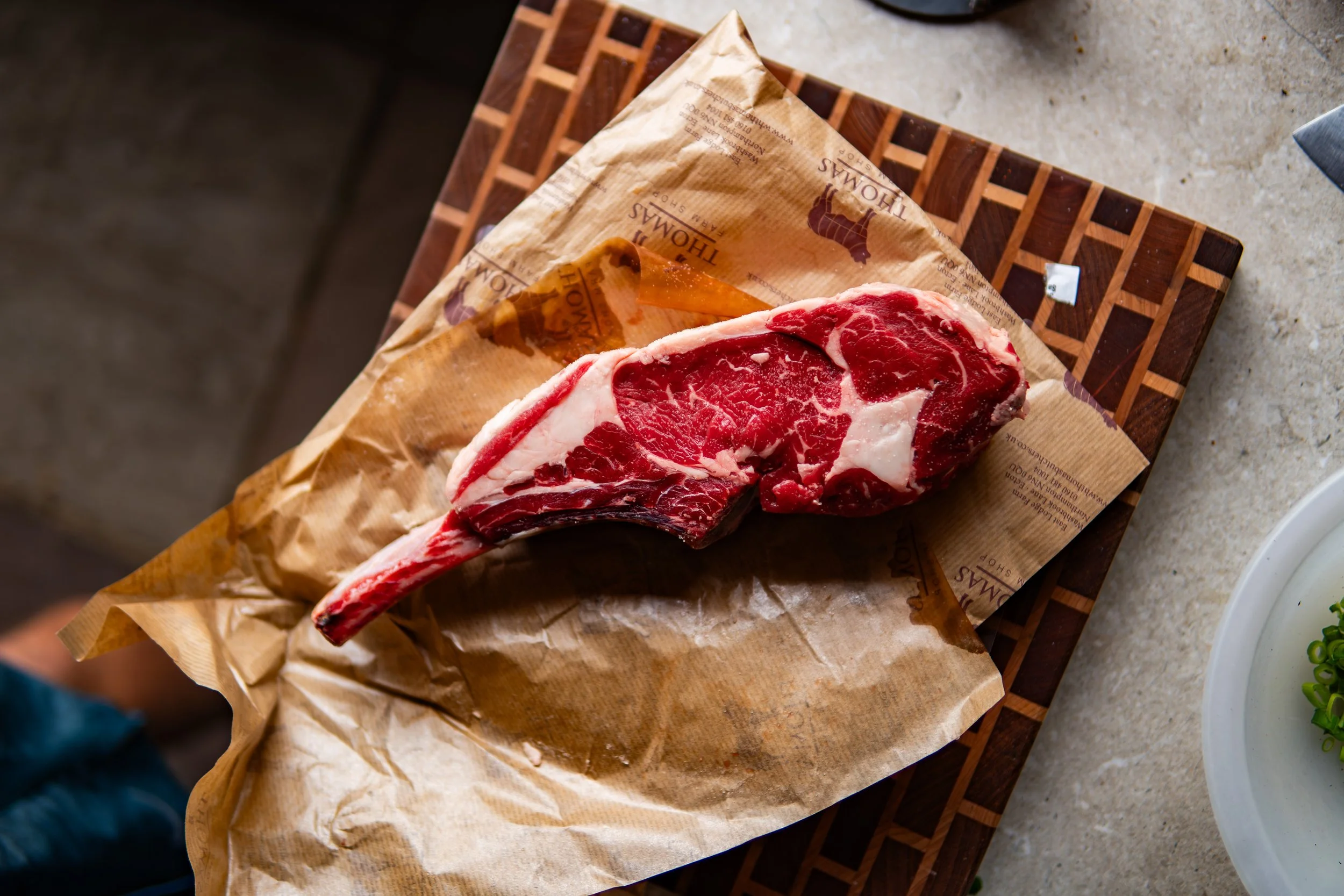 A raw ribeye steak on a wooden cutting board, wrapped partially in parchment paper, with a textured concrete surface nearby.