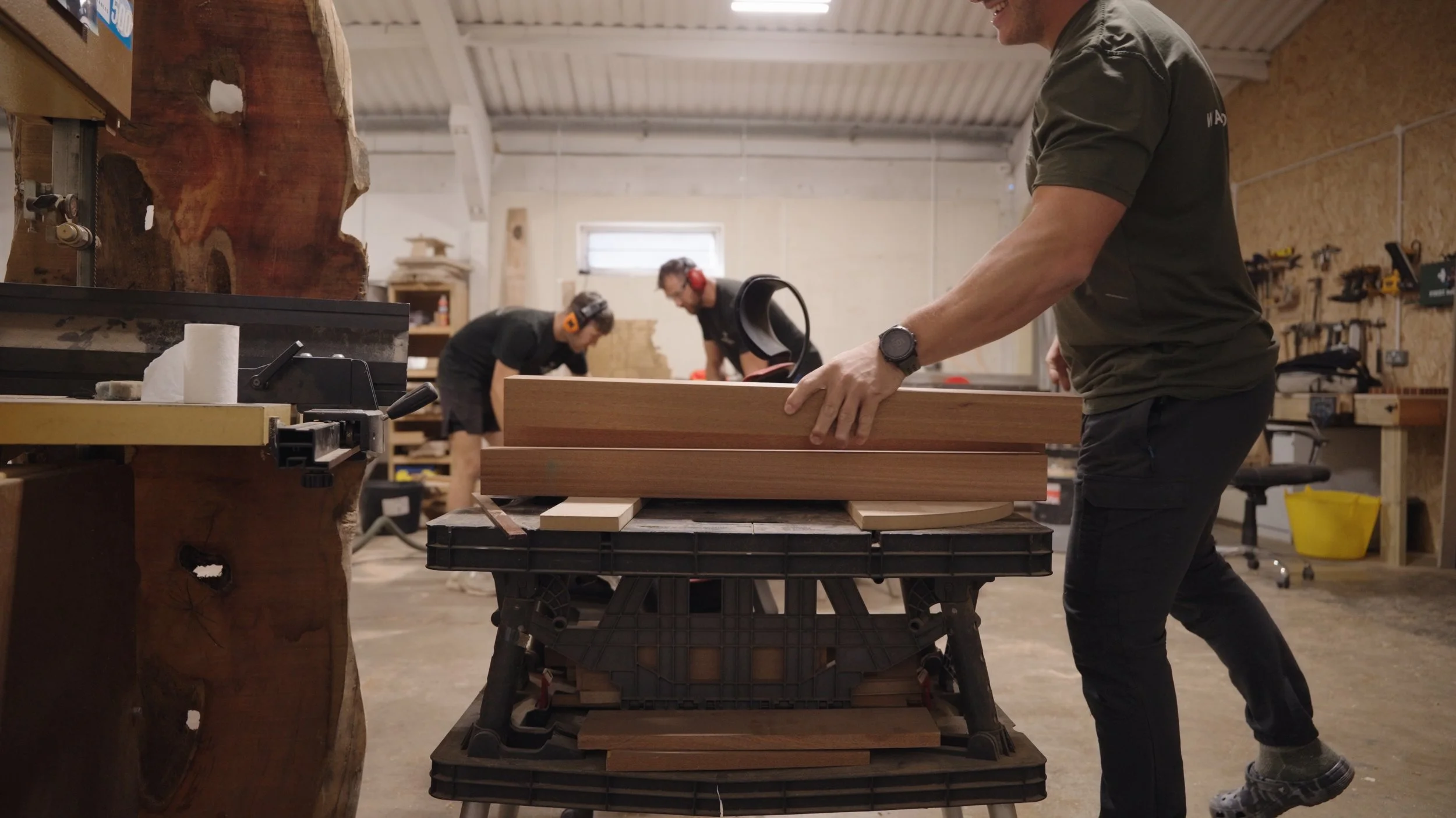 A person is moving a piece of wood in a woodworking shop with two other people working in the background, all wearing hearing protection.