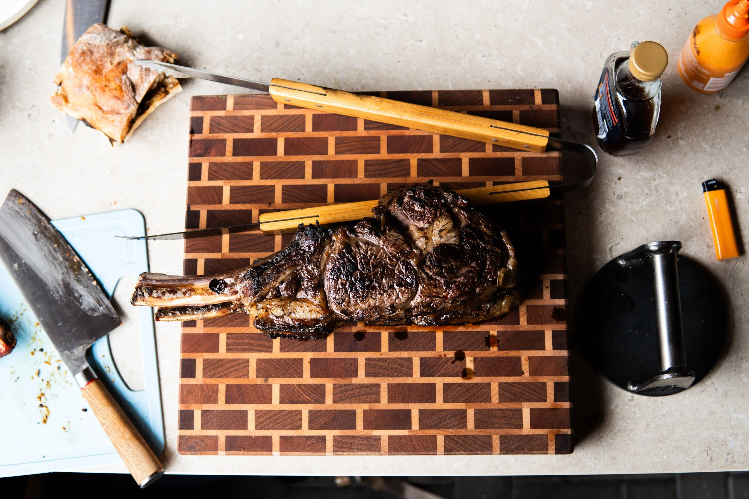 A cooked steak on a wooden cutting board with utensils, bottles of sauces, and a knife around it.