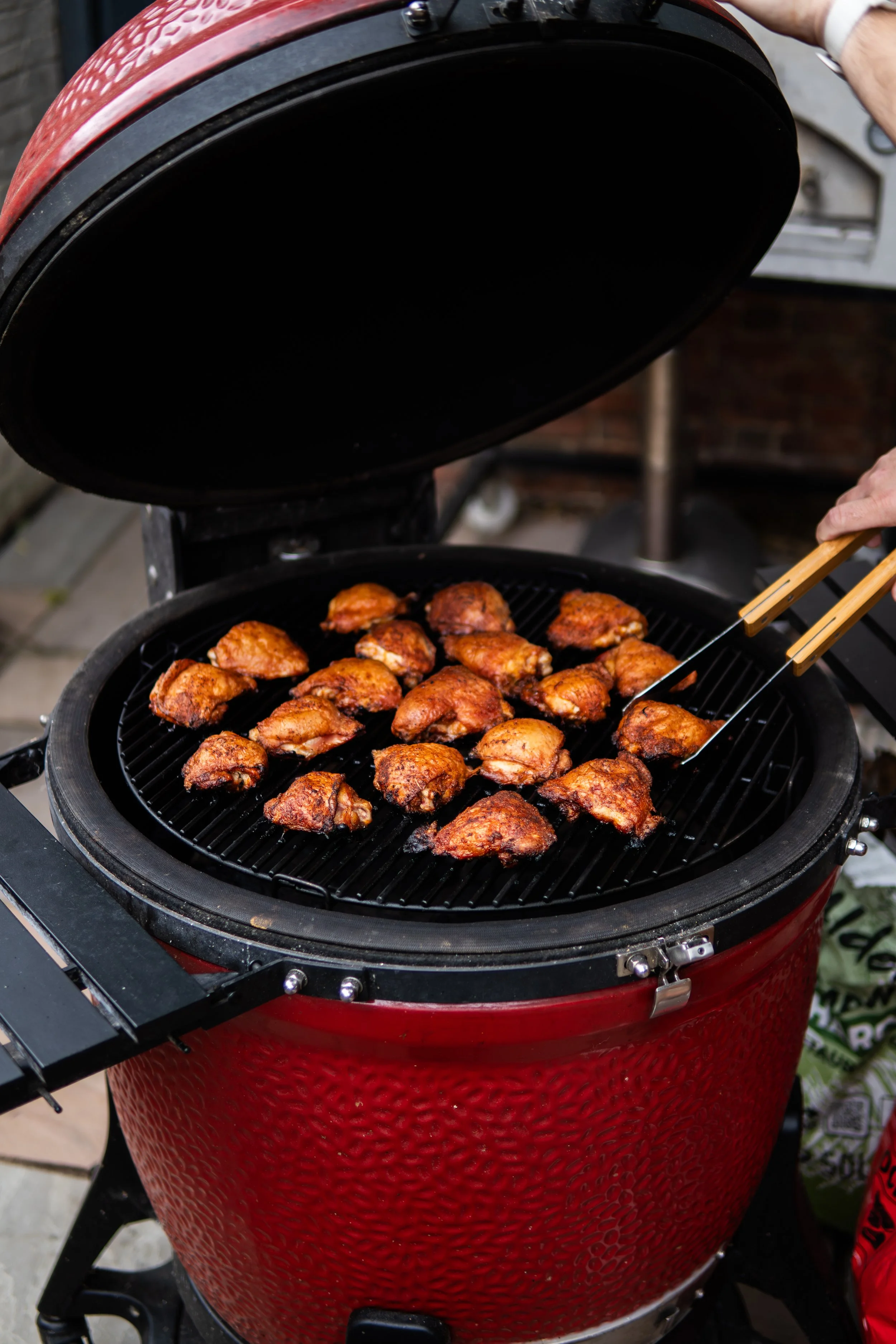 Barbecue grill with chicken drumsticks cooking on it, with someone using tongs to turn or check the meat.