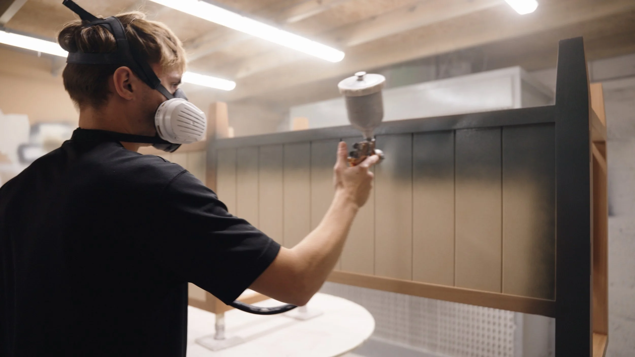 A person wearing a protective mask and ear protection is spray painting a piece of furniture in a workshop.