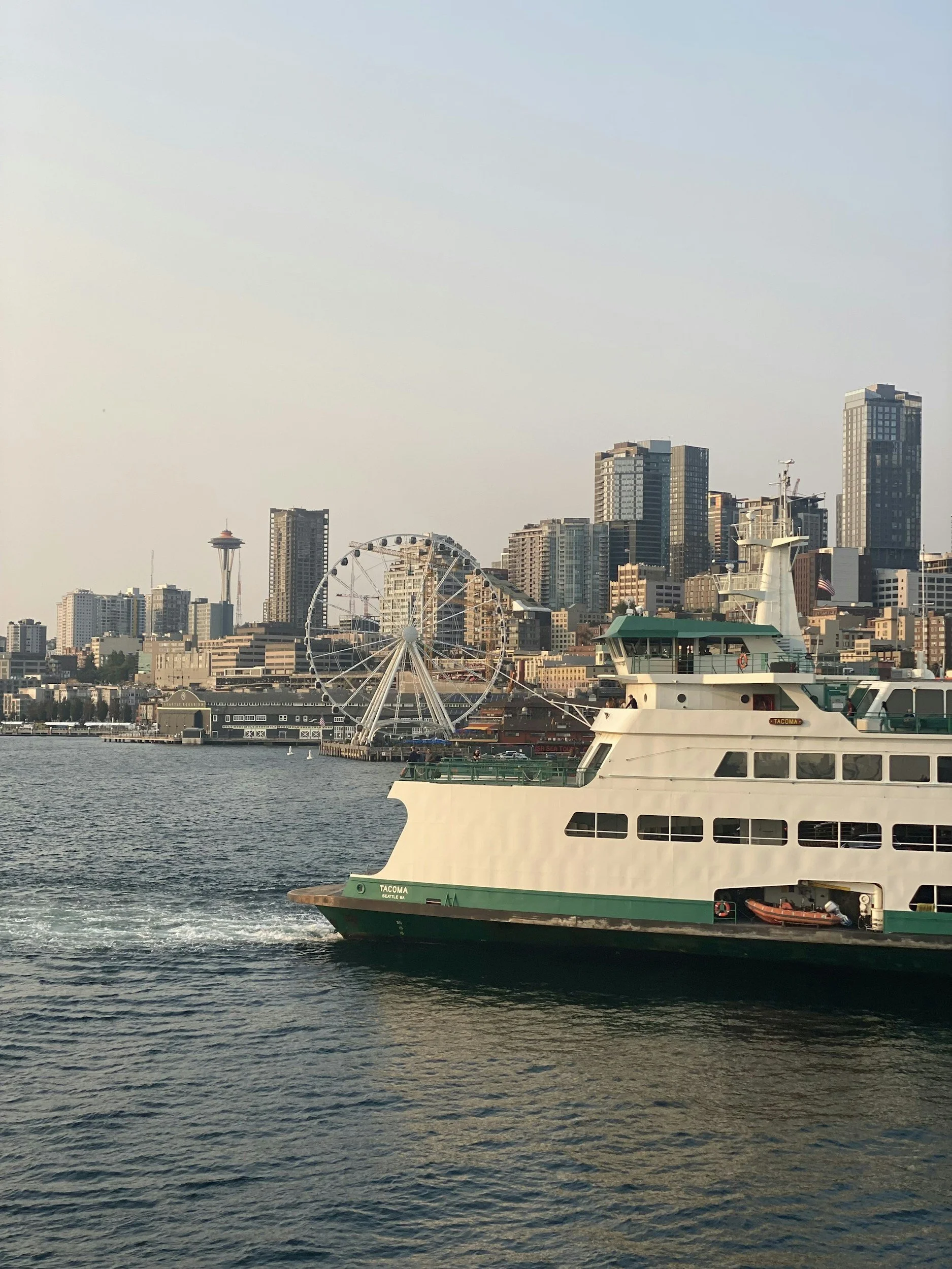 A ferry boat named Tacoma sailing on the water with the city skyline of Seattle, Washington, in the background, including the Space Needle and a large Ferris wheel.
