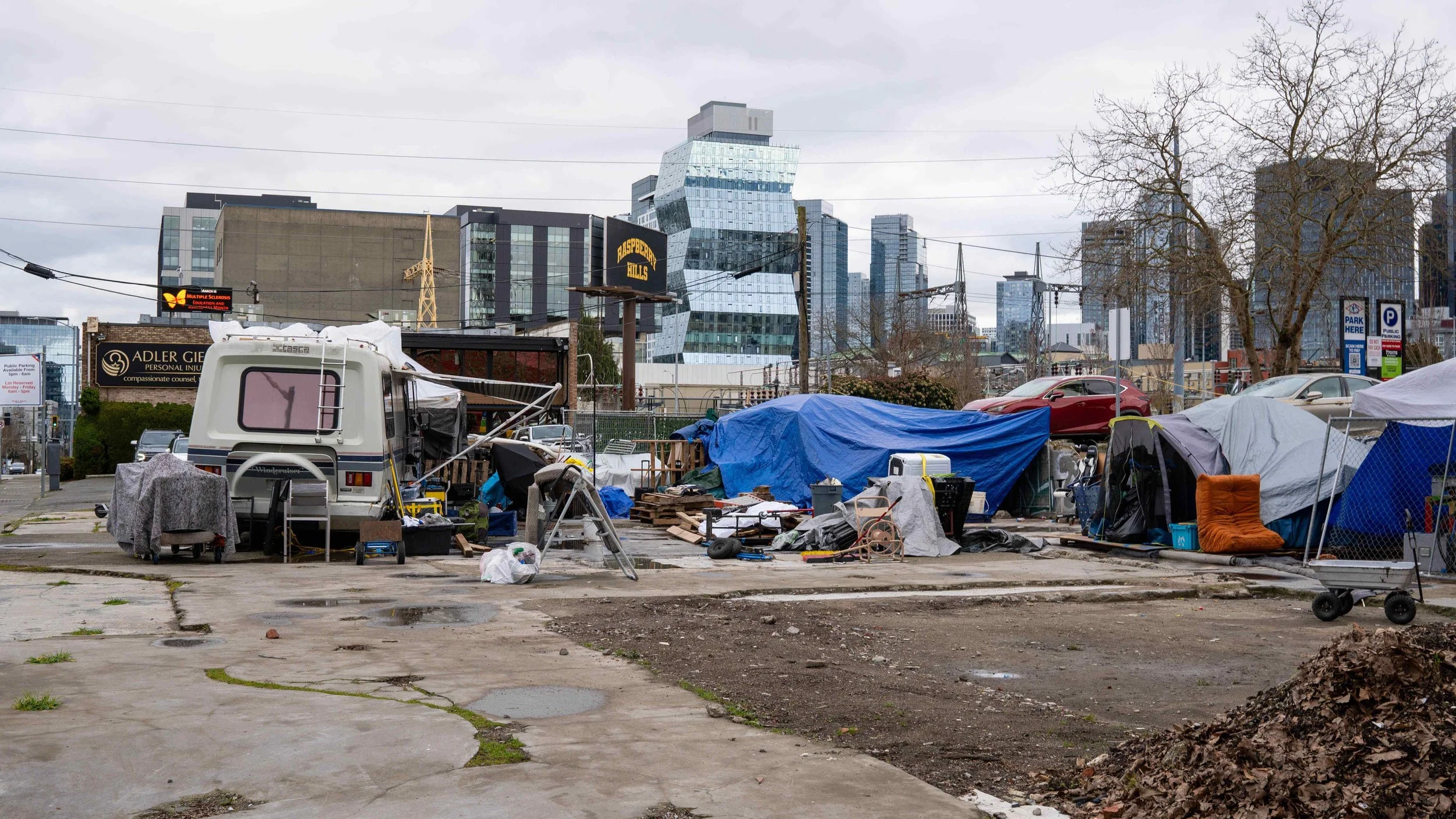 A large-scale RV and tent encampment on a paved lot with the South Lake Union tech skyline and 5th Ave North Garage in the background.