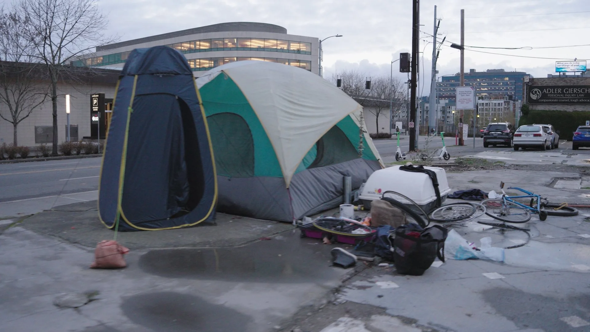 A small camping tent set up on a city sidewalk with scattered personal belongings around it, including bicycles, bags, and various items, with parked cars and city buildings in the background.