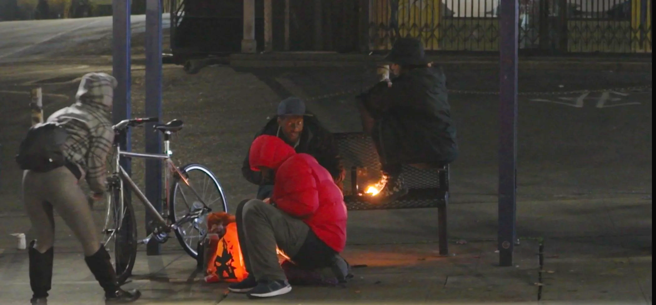 A group of four people at night around a small fire in an urban setting. One person is sitting on the ground in a red jacket, another is squatting nearby, and two are sitting on a bench, one wearing a black hat and the other in a black hoodie. A bicycle and a hooded figure are also visible.