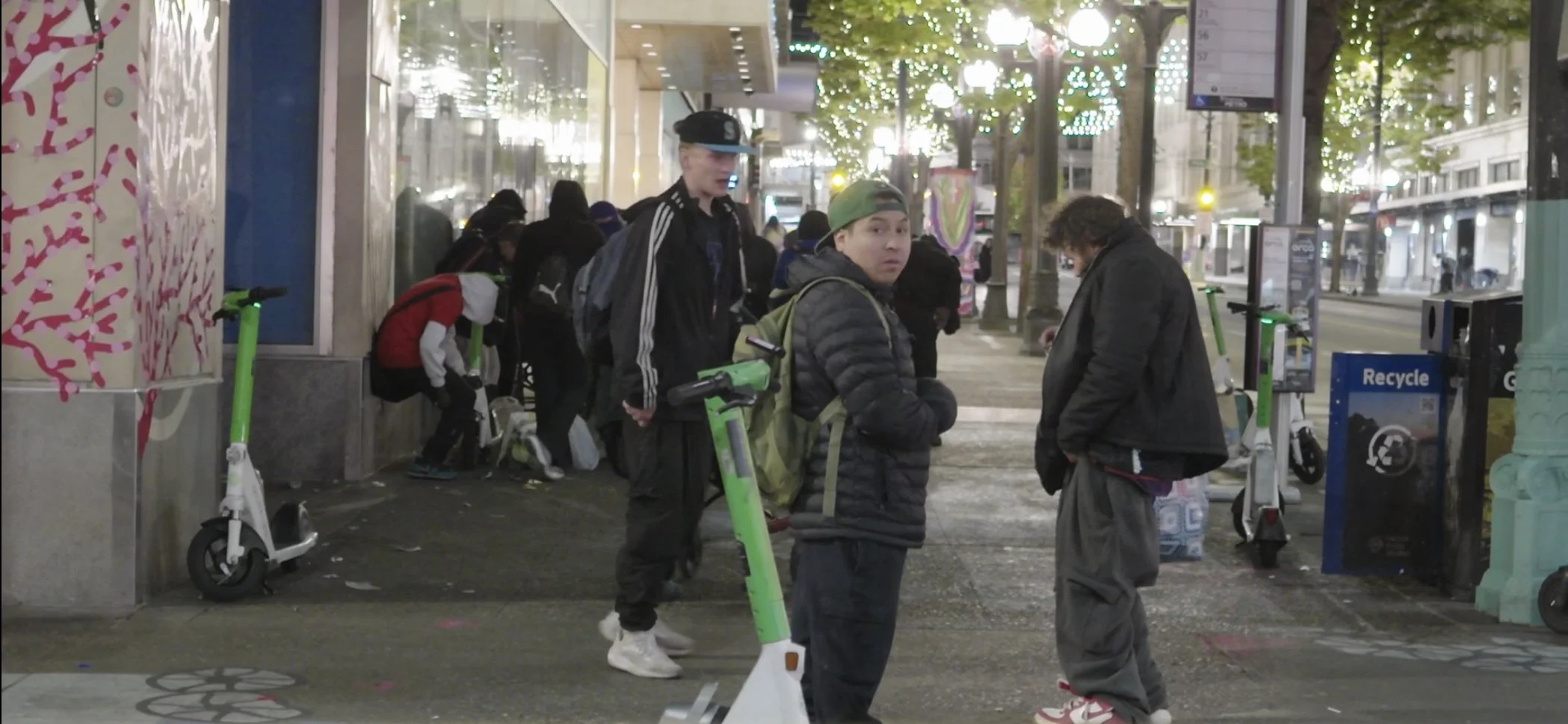 A group of people on a city sidewalk at night, some with backpacks, near illuminated trees and storefronts, with green electric scooters parked along the curb.