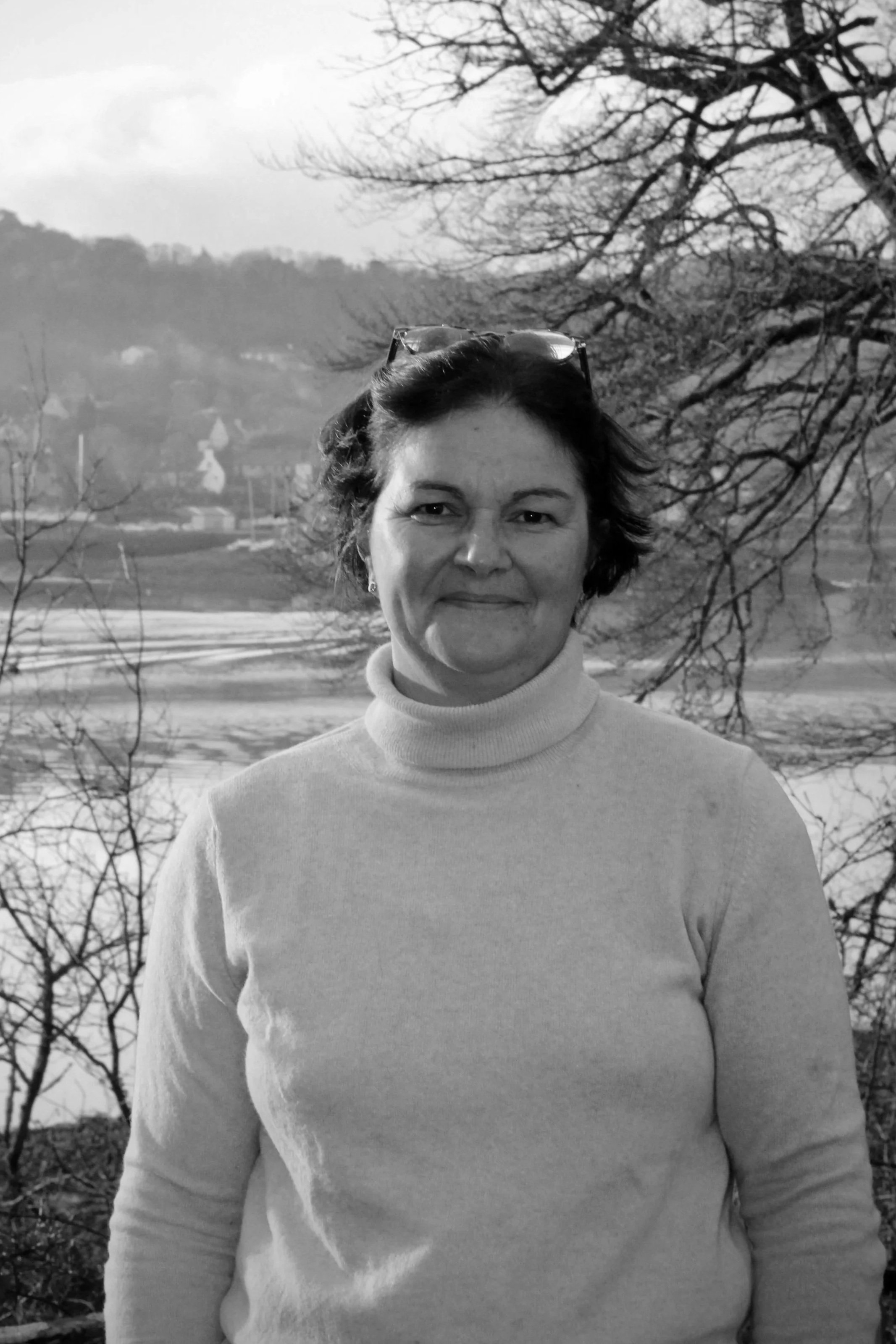 Une femme avec des lunettes posées sur sa tête, portant un pull à col roulé, en extérieur avec un paysage de champs et d'arbres en arrière-plan, en noir et blanc.