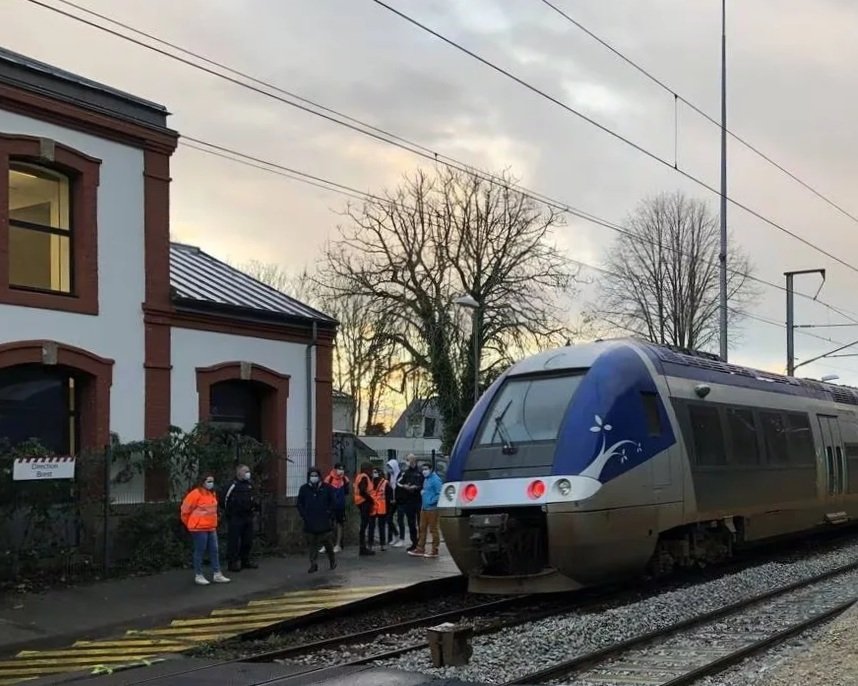Un groupe de personnes attendant le train à une station dans une région urbaine avec un bâtiment en arrière-plan, sous un ciel nuageux, et un train moderne arrêté sur la voie