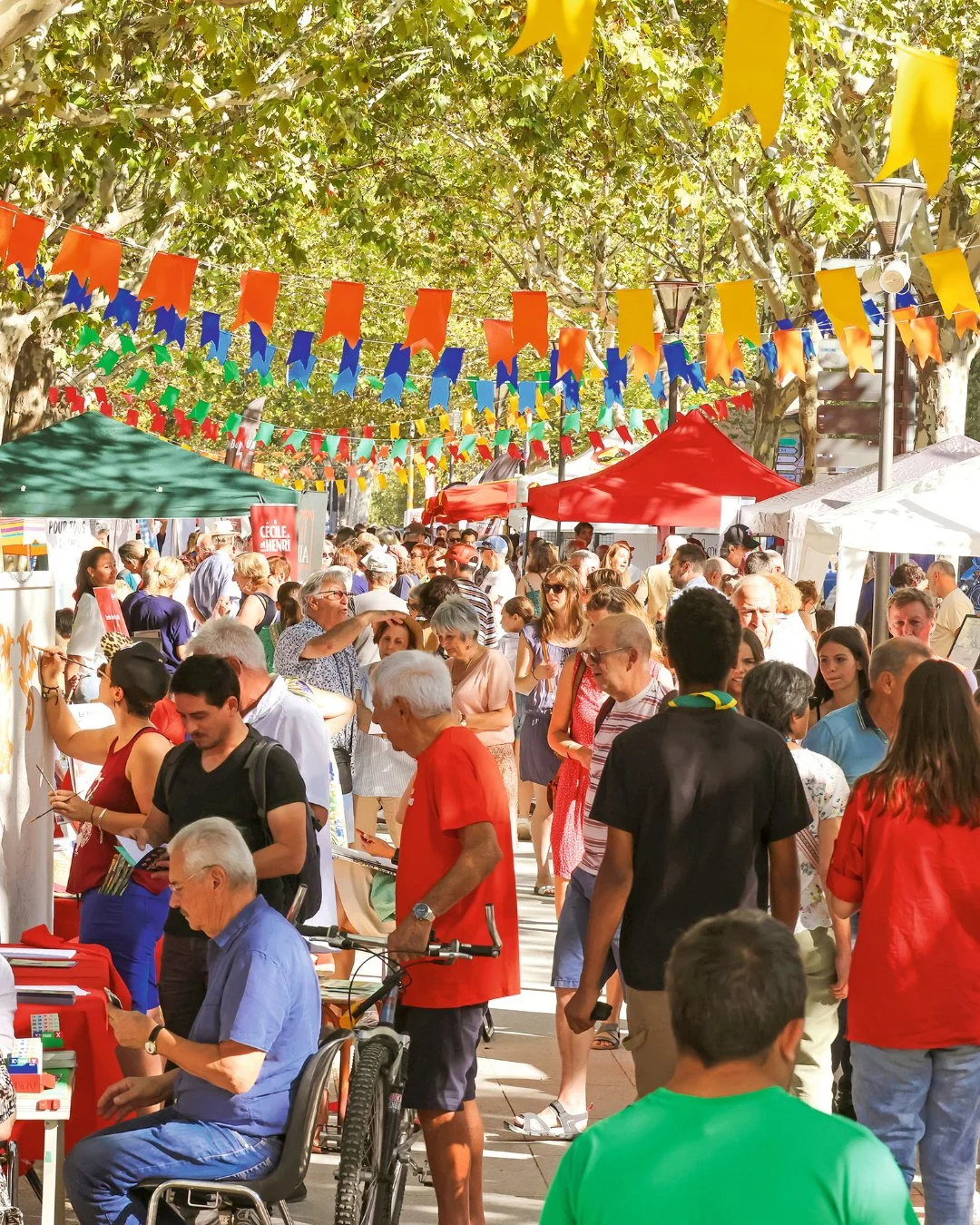 Une foule de personnes lors d'une fête en plein air avec des banderoles colorées suspendues au-dessus, plusieurs stands avec tentes de différentes couleurs, arbres en fond, ambiance festive et animée.