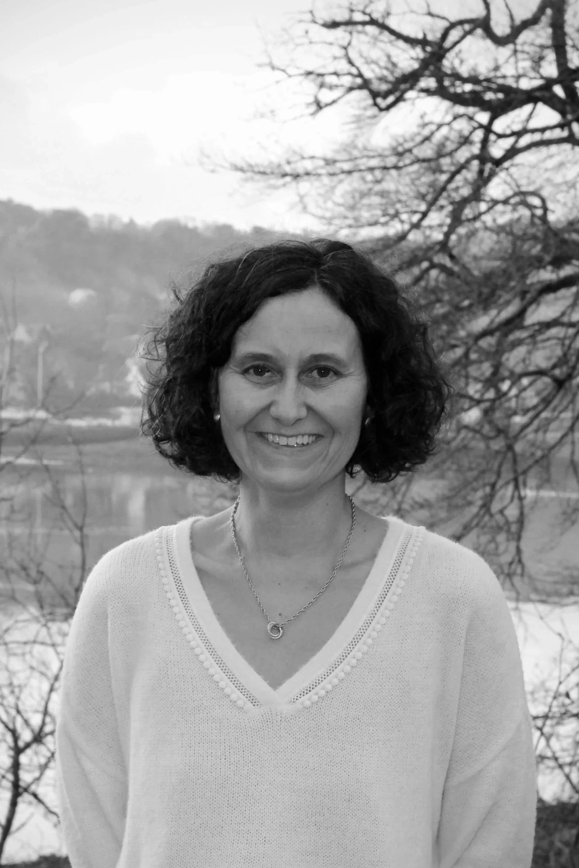 Portrait en noir et blanc d'une femme souriante avec des cheveux bouclés, portant un pull blanc, un collier et des boucles d'oreilles, avec un arbre sans feuilles et un lac en arrière-plan.