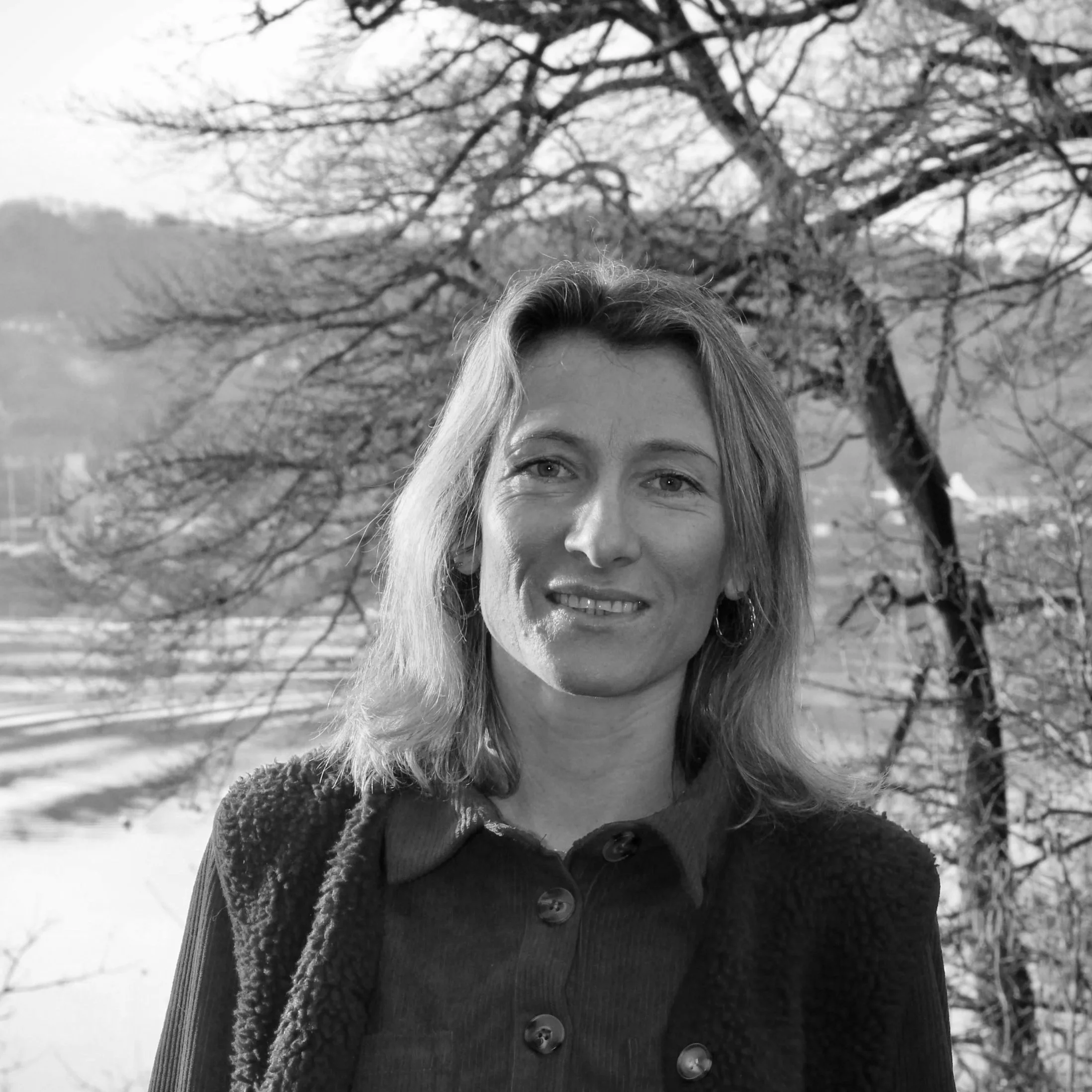 Une femme souriante avec des cheveux mi-longs, portant un manteau, en extérieur avec un arbre dénudé en arrière-plan.