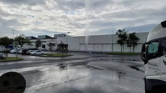 vue d'une zone de parking devant un bâtiment moderne sous un ciel nuageux, avec des voitures et un camion sur le côté droit.