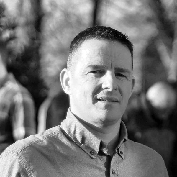 Un homme adulte regarde la caméra, portant une chemise à col, avec un arrière-plan flou d'arbres et de personnes. L'image est en noir et blanc.