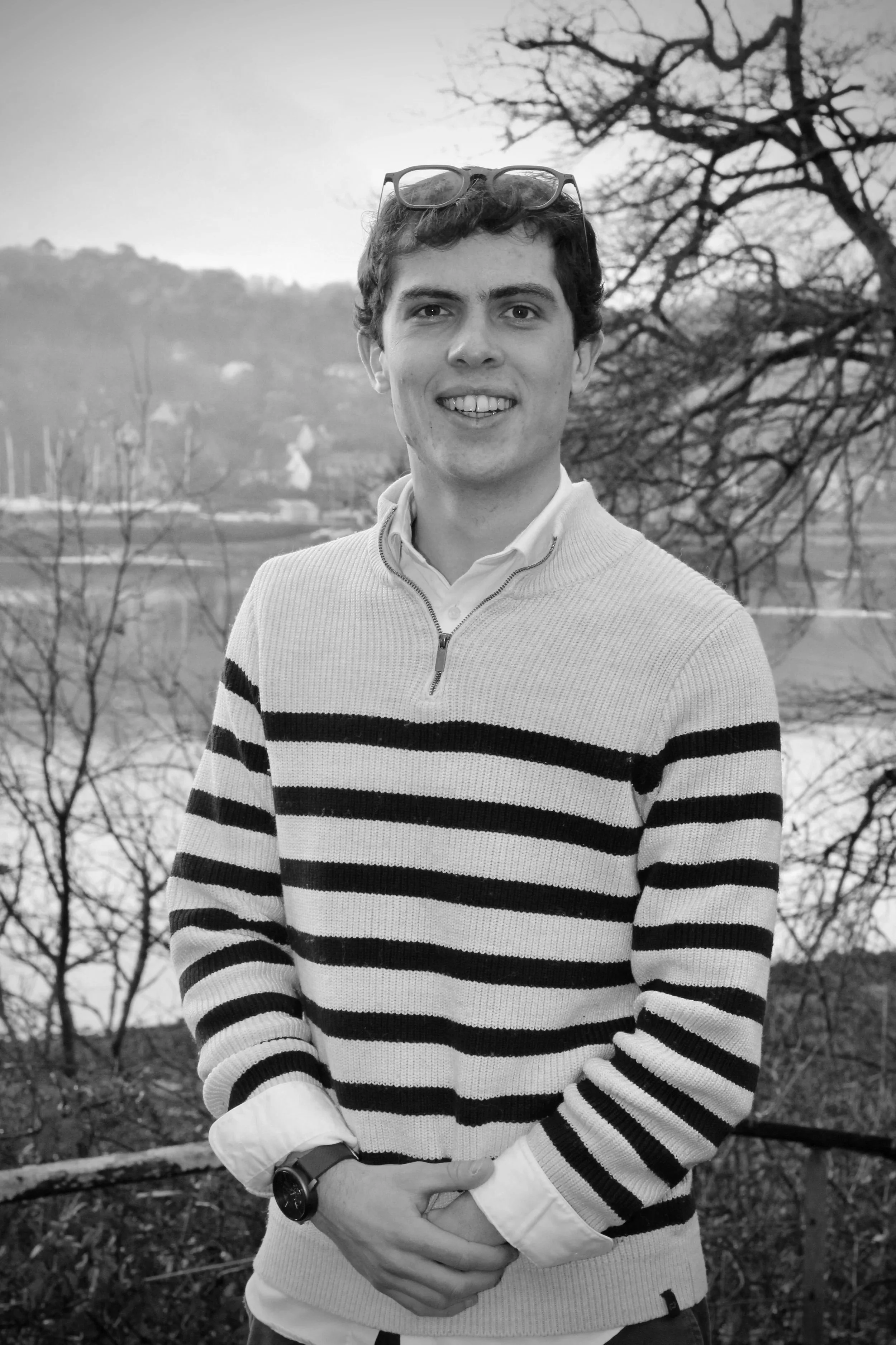 Jeune homme souriant portant un pull rayé noir et blanc, lunettes sur la tête, tenant ses mains devant lui, avec un paysage naturel en arrière-plan, en noir et blanc.